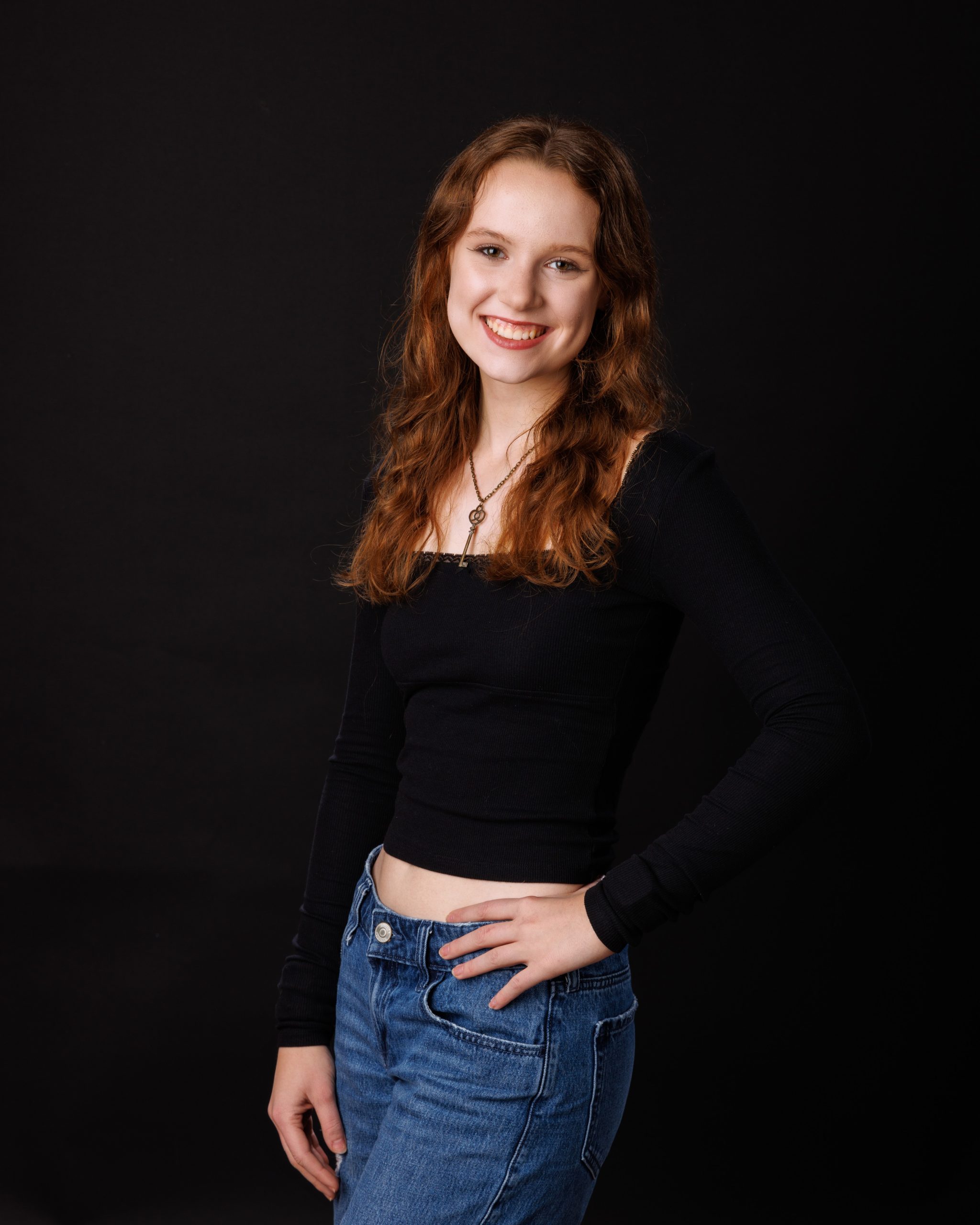 Female student in black top and jeans posing for senior portrait with long hair against dark background