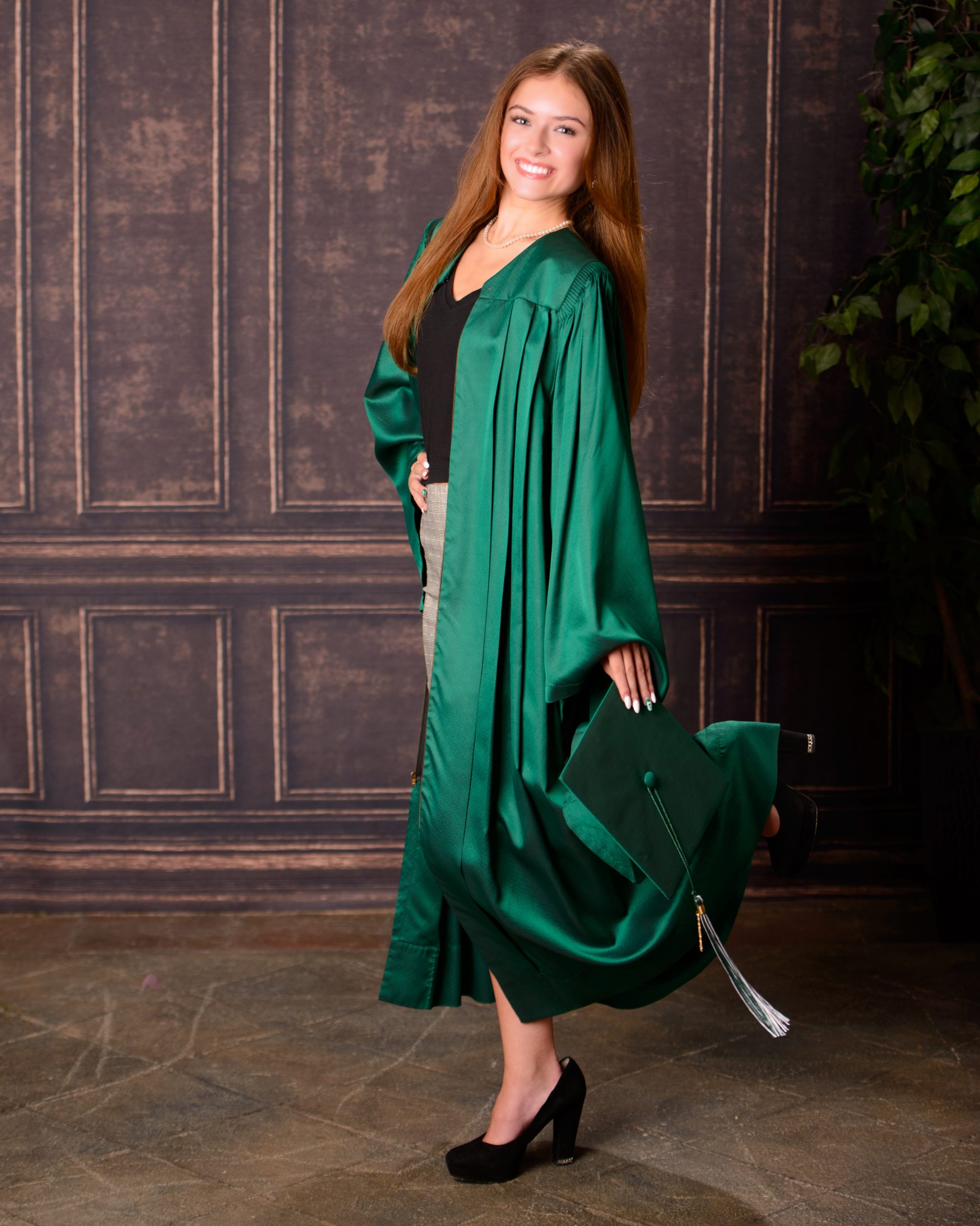 Senior graduate posing with cap held up, wearing green gown and black heels