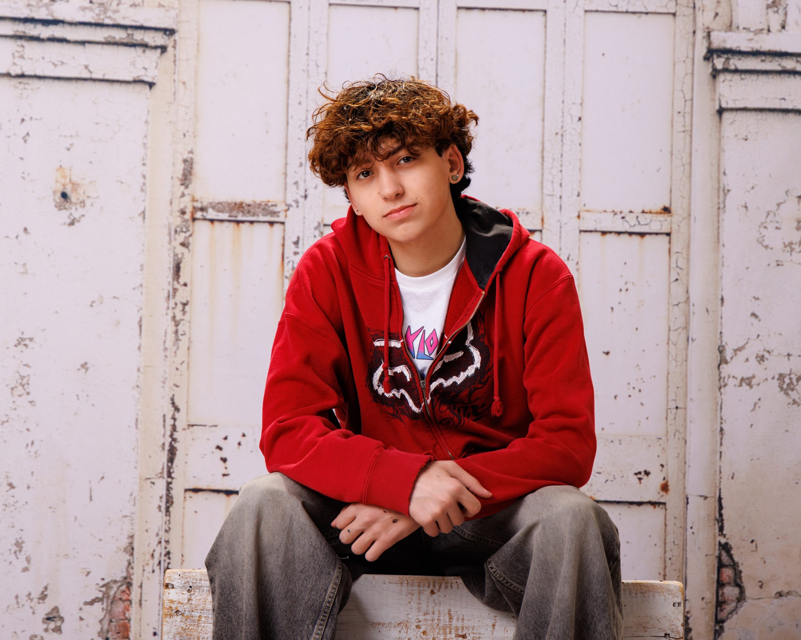 Diego seated on wooden bench in red hoodie, white tee, weathered door background