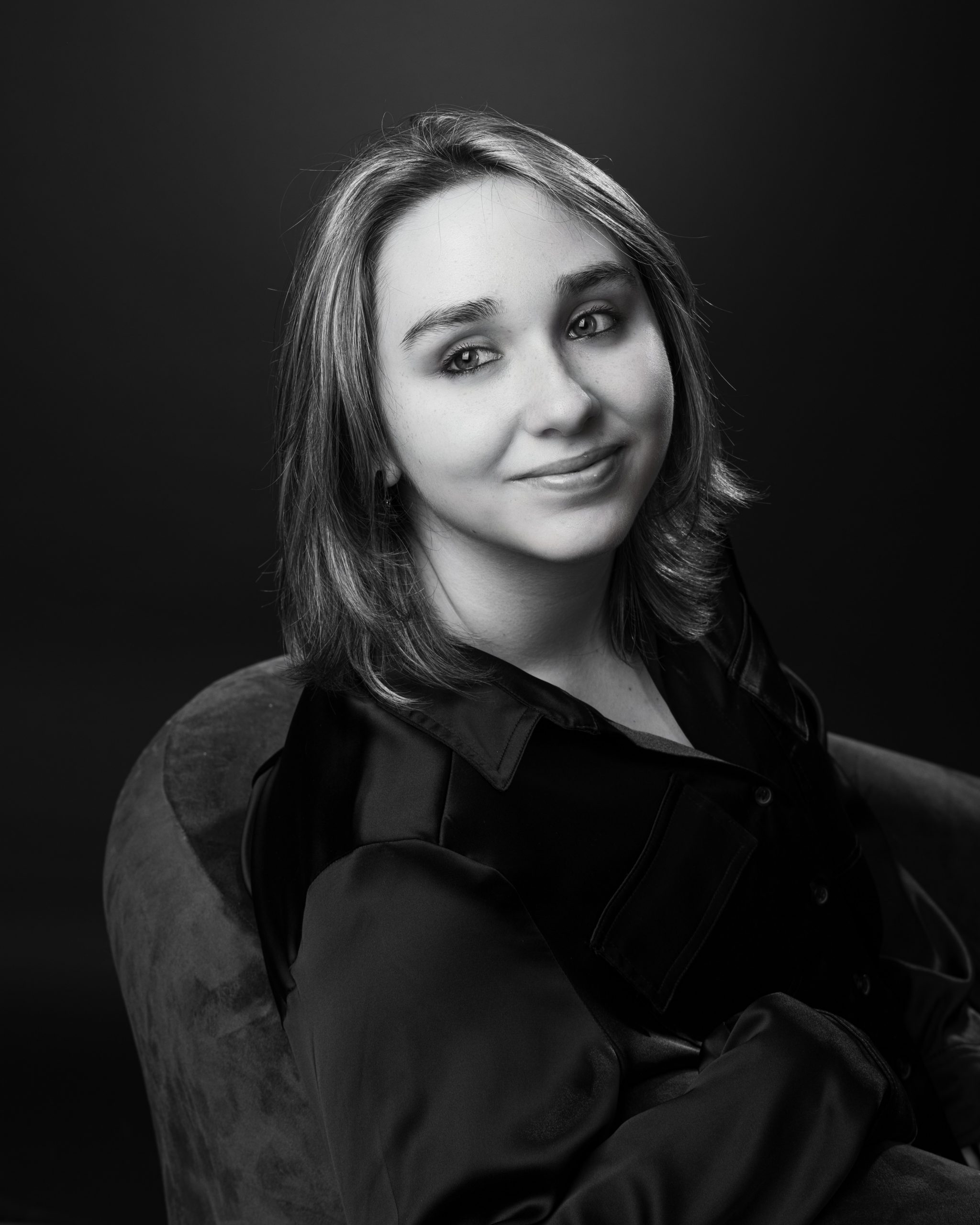 Senior portrait of Emma in black satin blouse seated on brown chair against dark backdrop