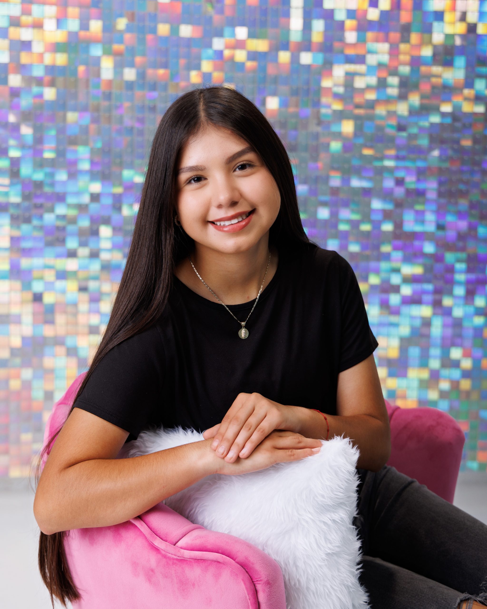 Senior girl Anahi posing on pink chair with white fluffy pillow against glitter pixel wall background