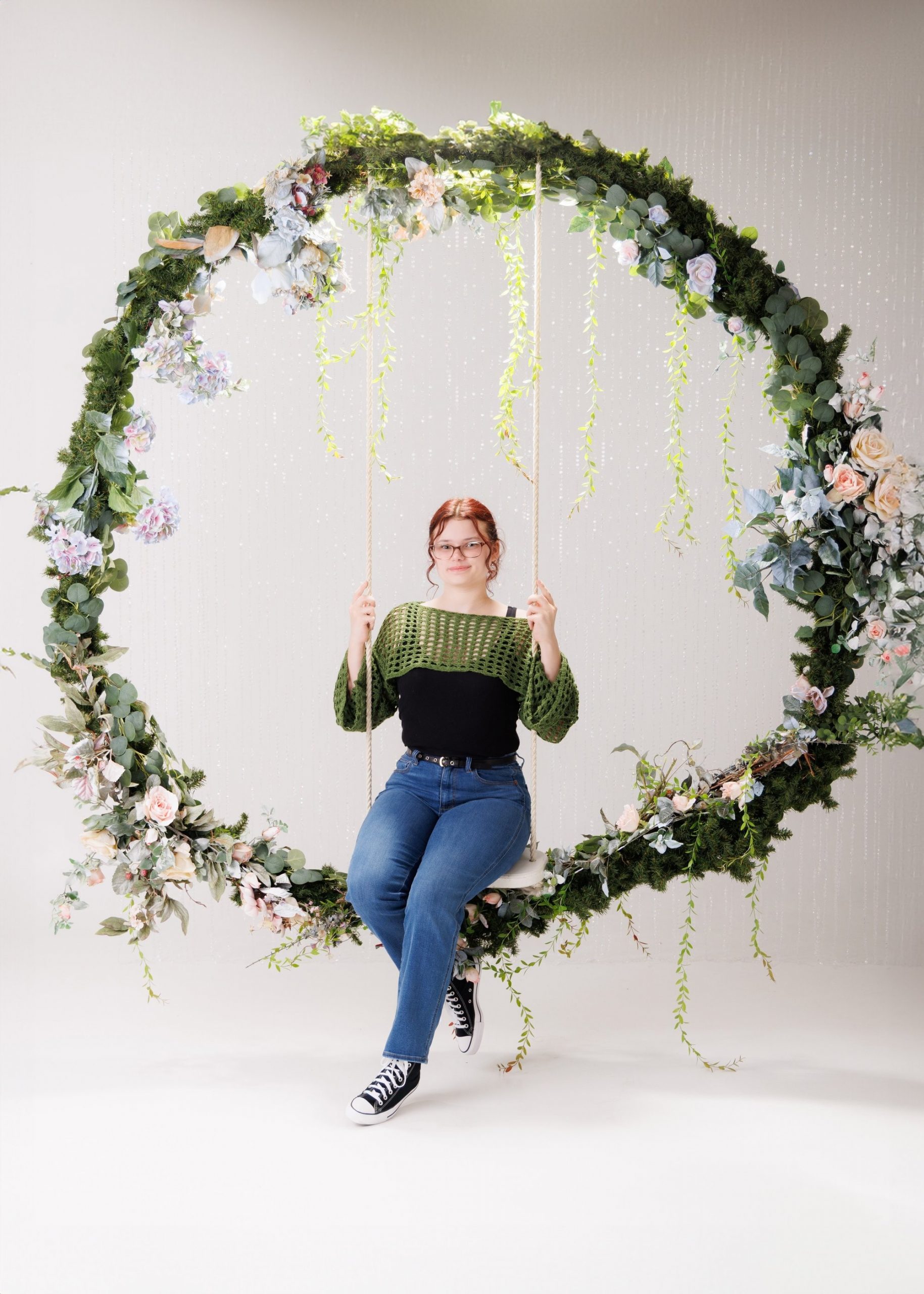 Grace posing on a swing with green crochet shawl and vibrant flower arrangements in the background