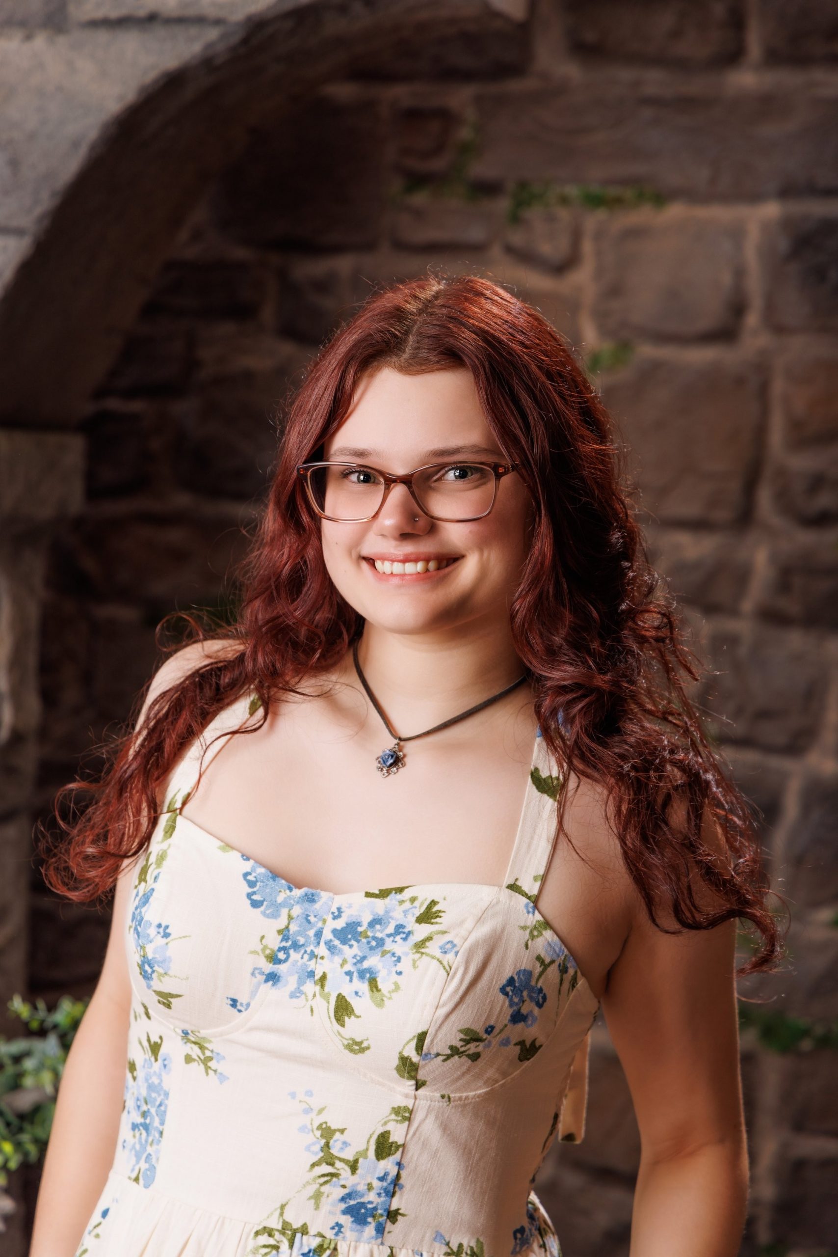 Senior portrait of Grace wearing a blue and green floral dress against a stone wall backdrop