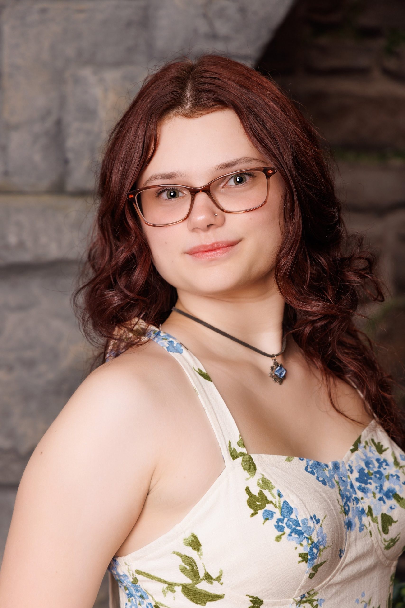 Senior portrait of Grace wearing a blue and green floral dress against a stone wall backdrop