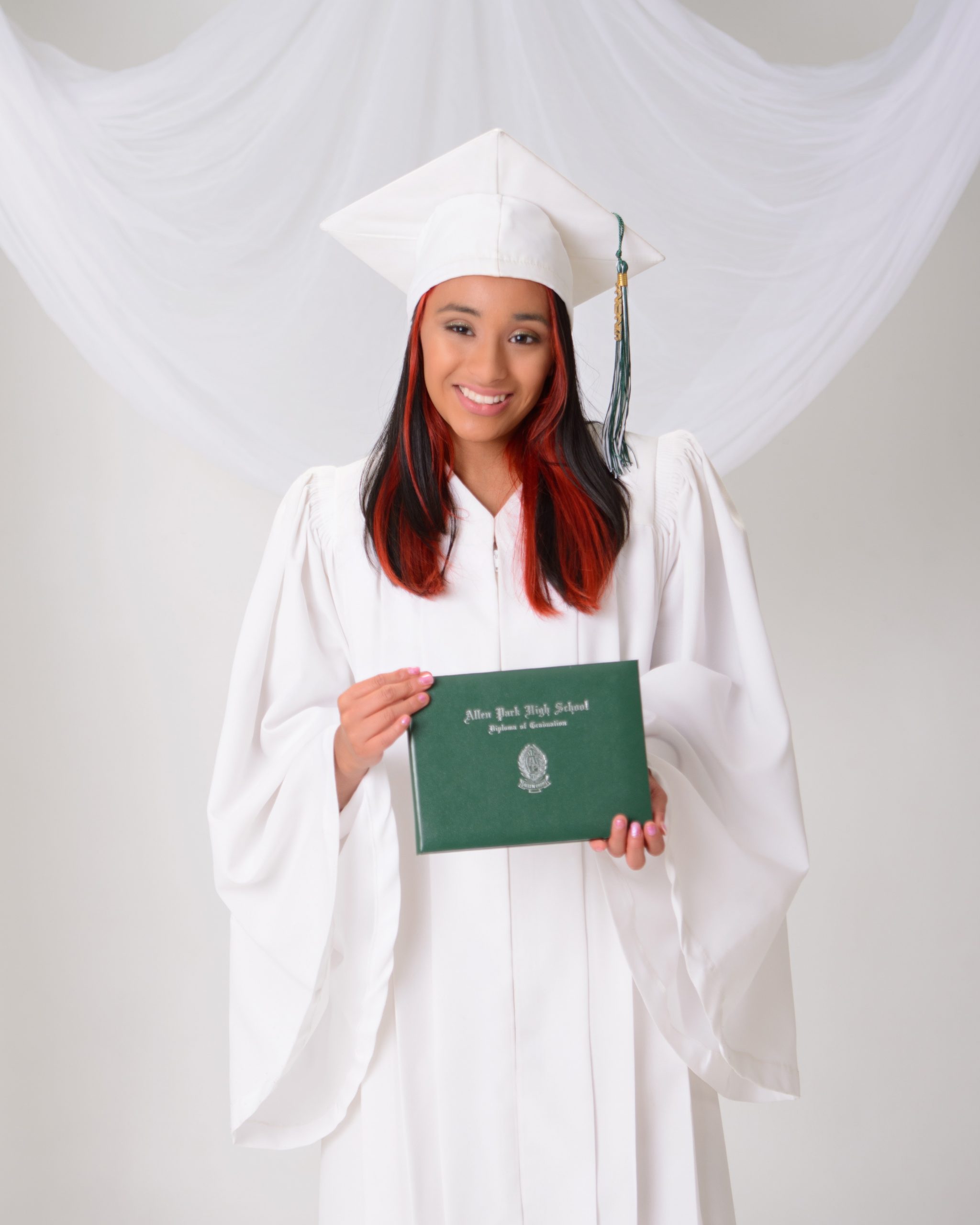 A graduate in a white cap and gown holds an Allen Park High School diploma, smiling at the camera.
