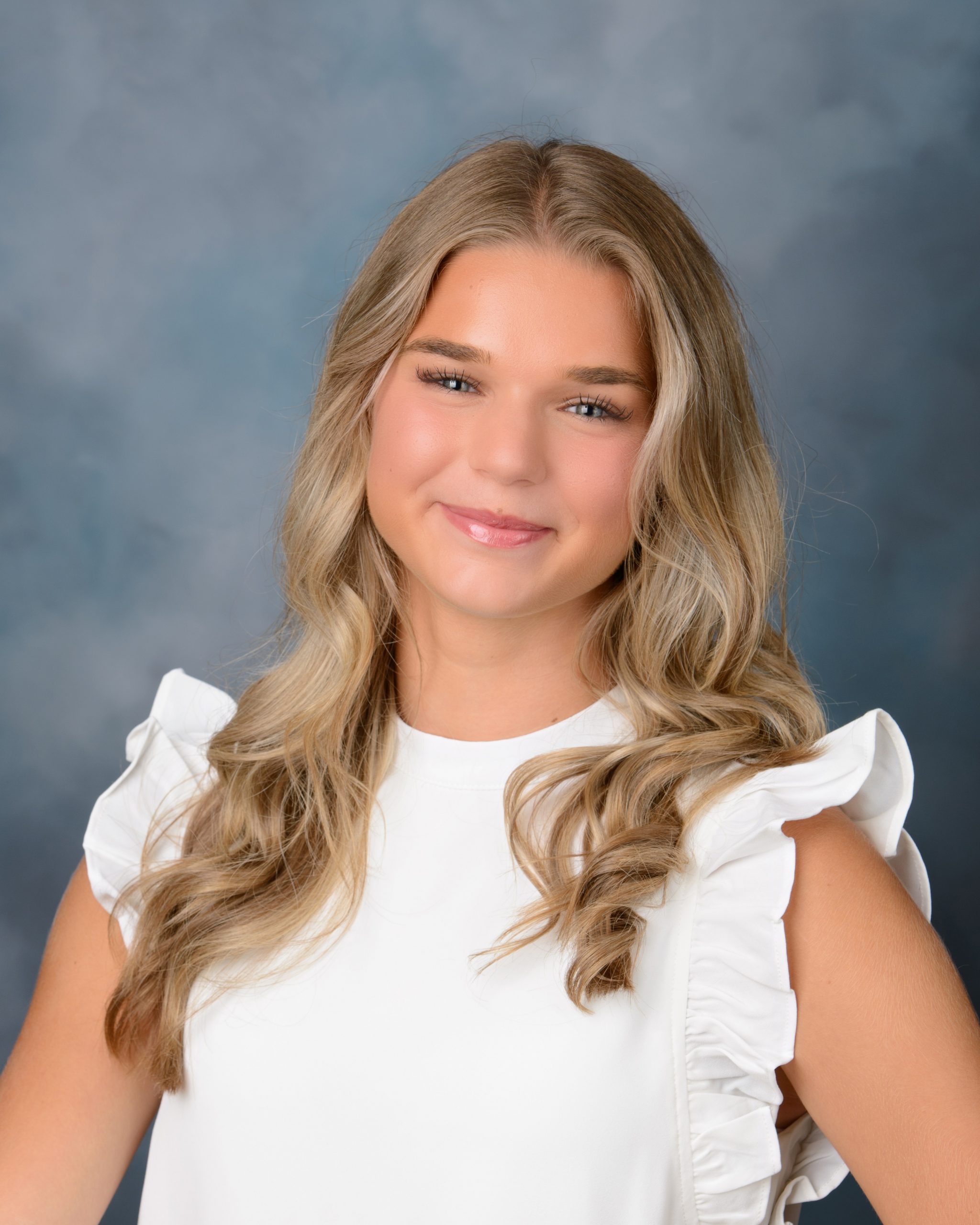 Senior portrait of Morgan in white ruffled top against blue studio backdrop