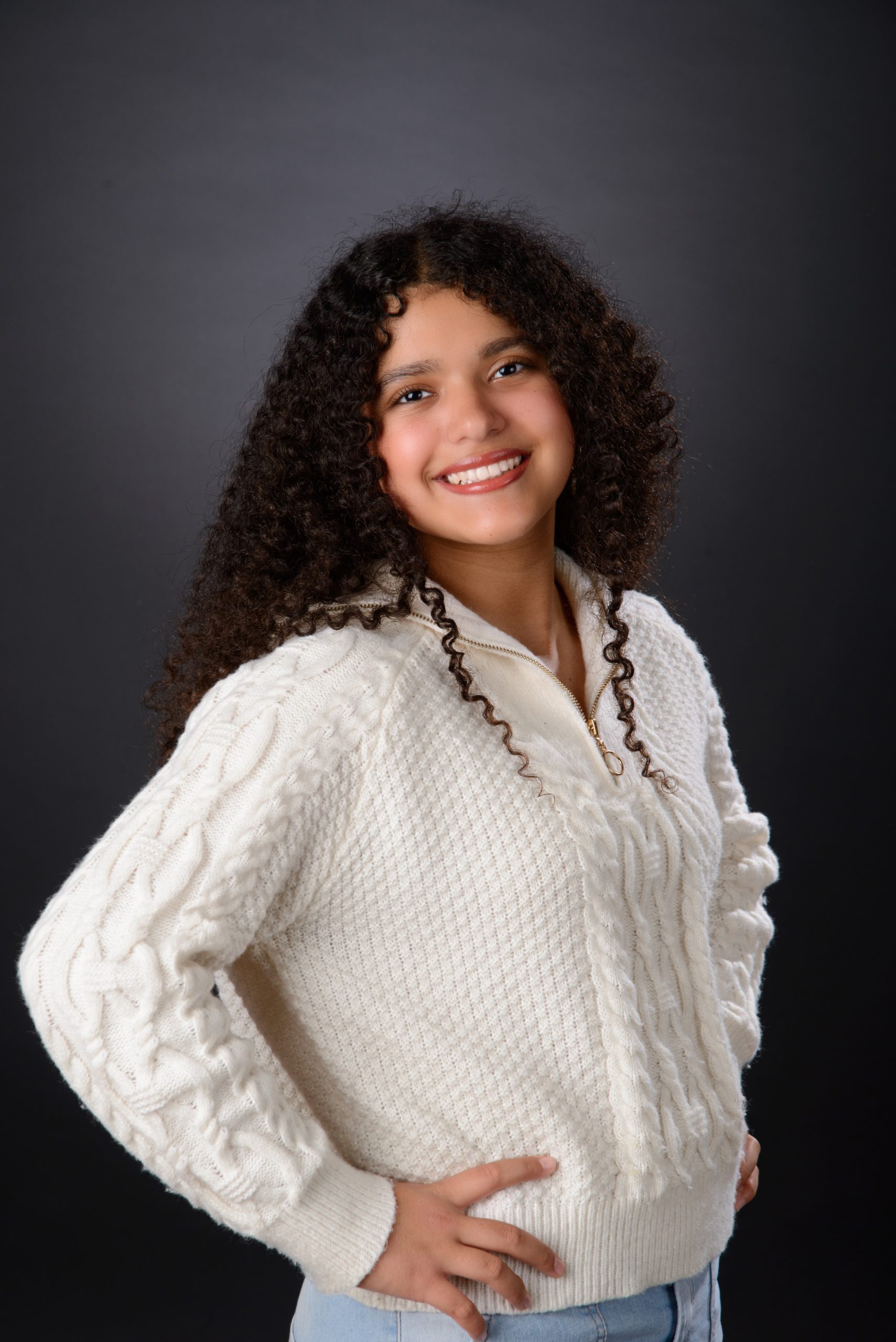 Class of 2025 senior in white sweater poses with hands on hips against dark backdrop