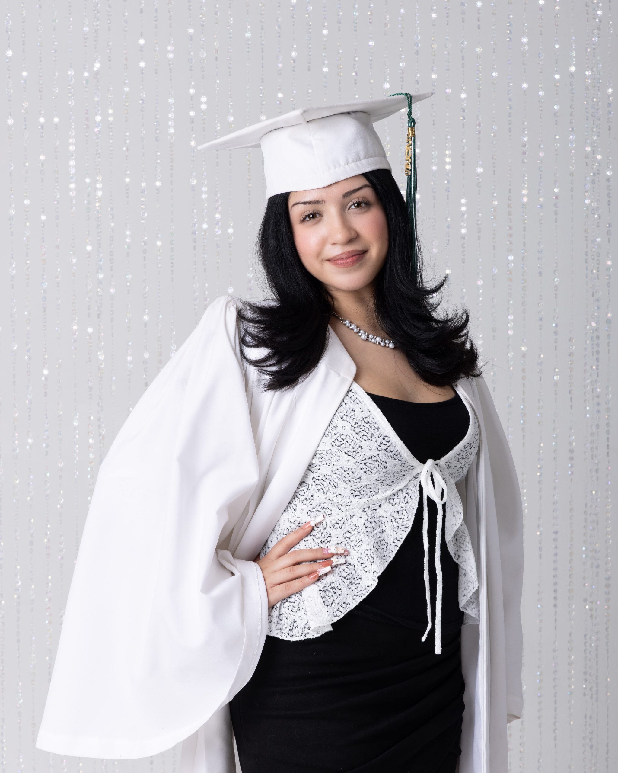 Graduation-ready senior photo featuring a student in white cap, lace cardigan, and black dress against light pink backdrop