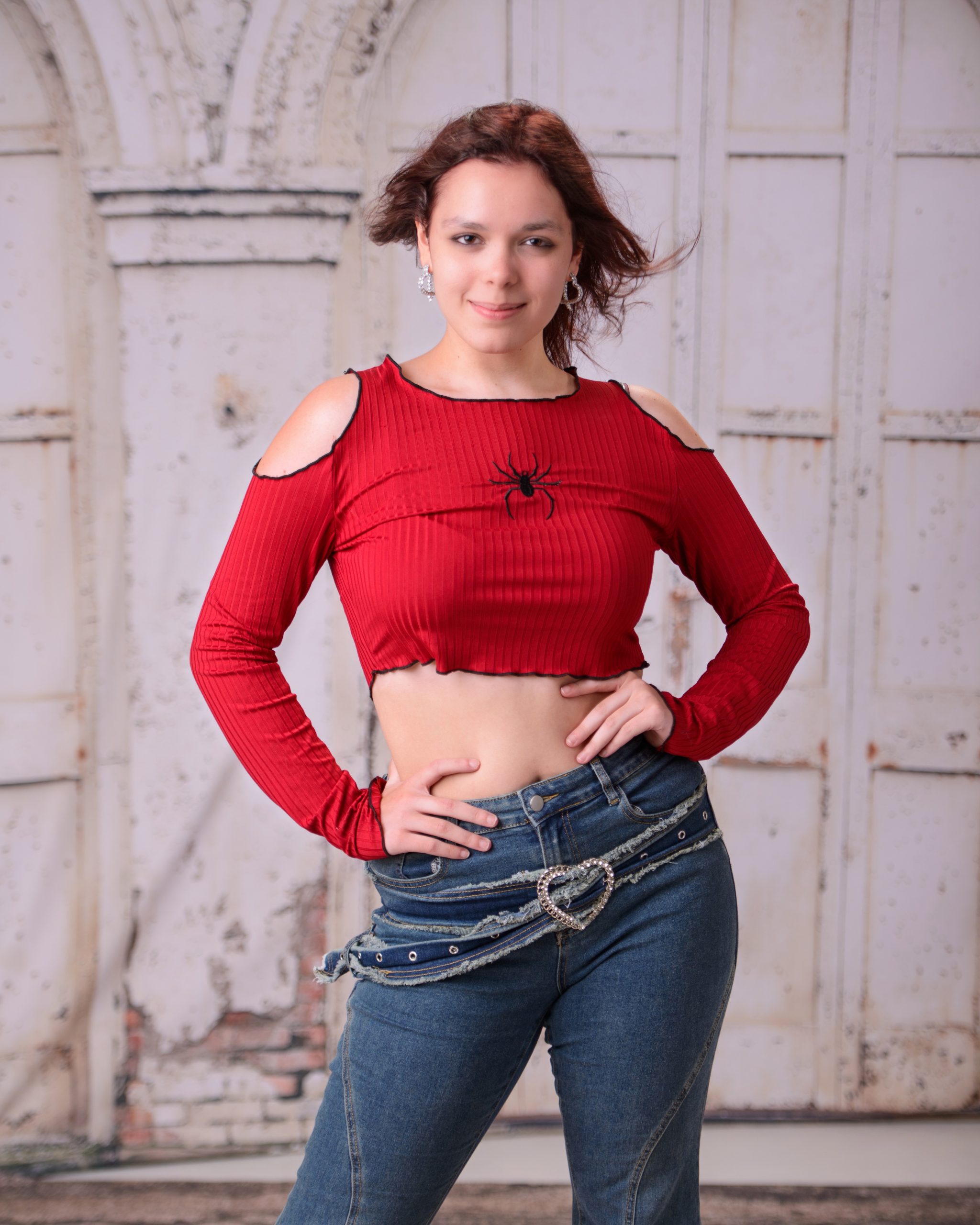 senior portrait of woman in red crop top with spider design and denim jeans posing against white door backdrop