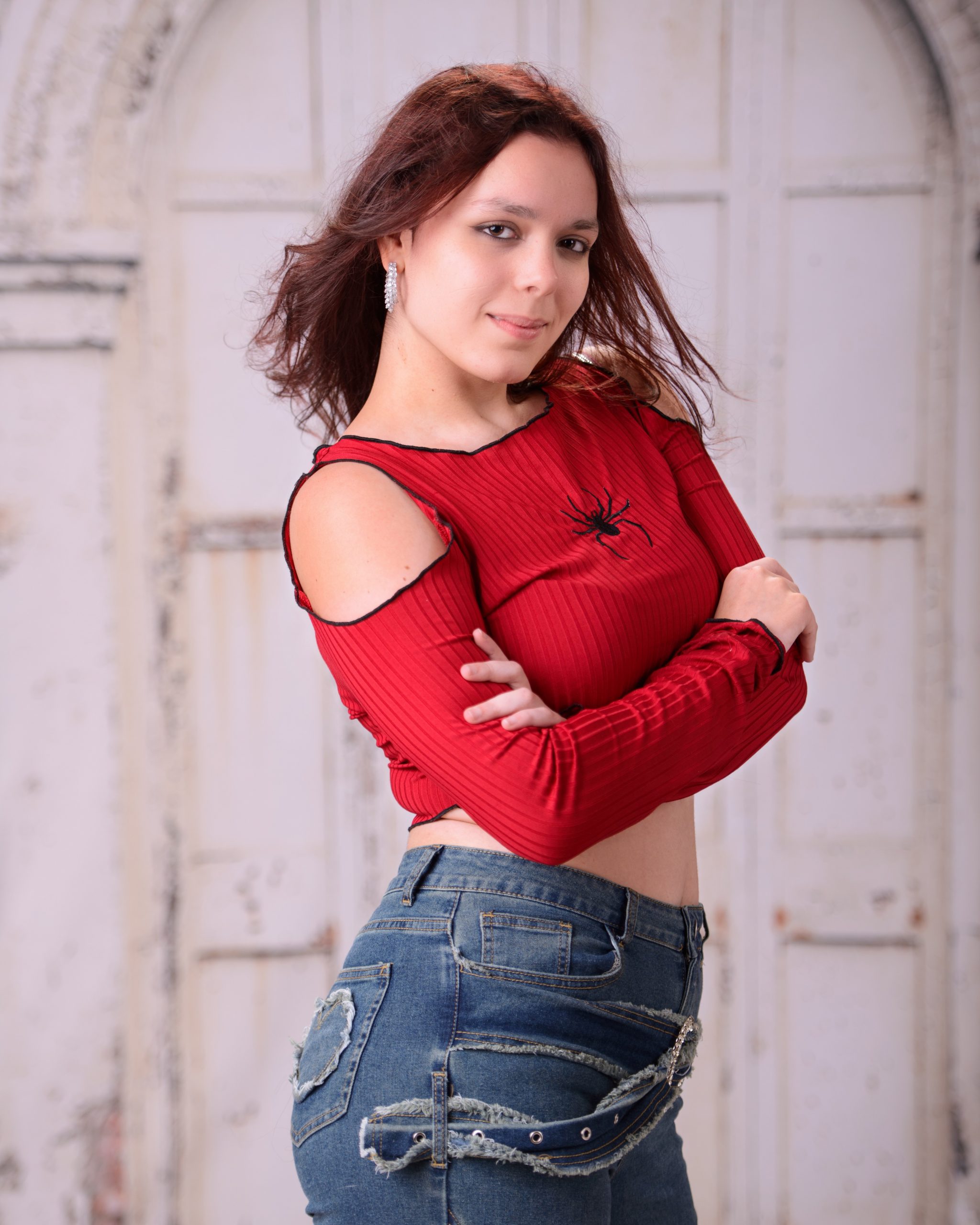 confident senior photo featuring a woman in red off-shoulder top and heart belt detail standing before weathered white doors