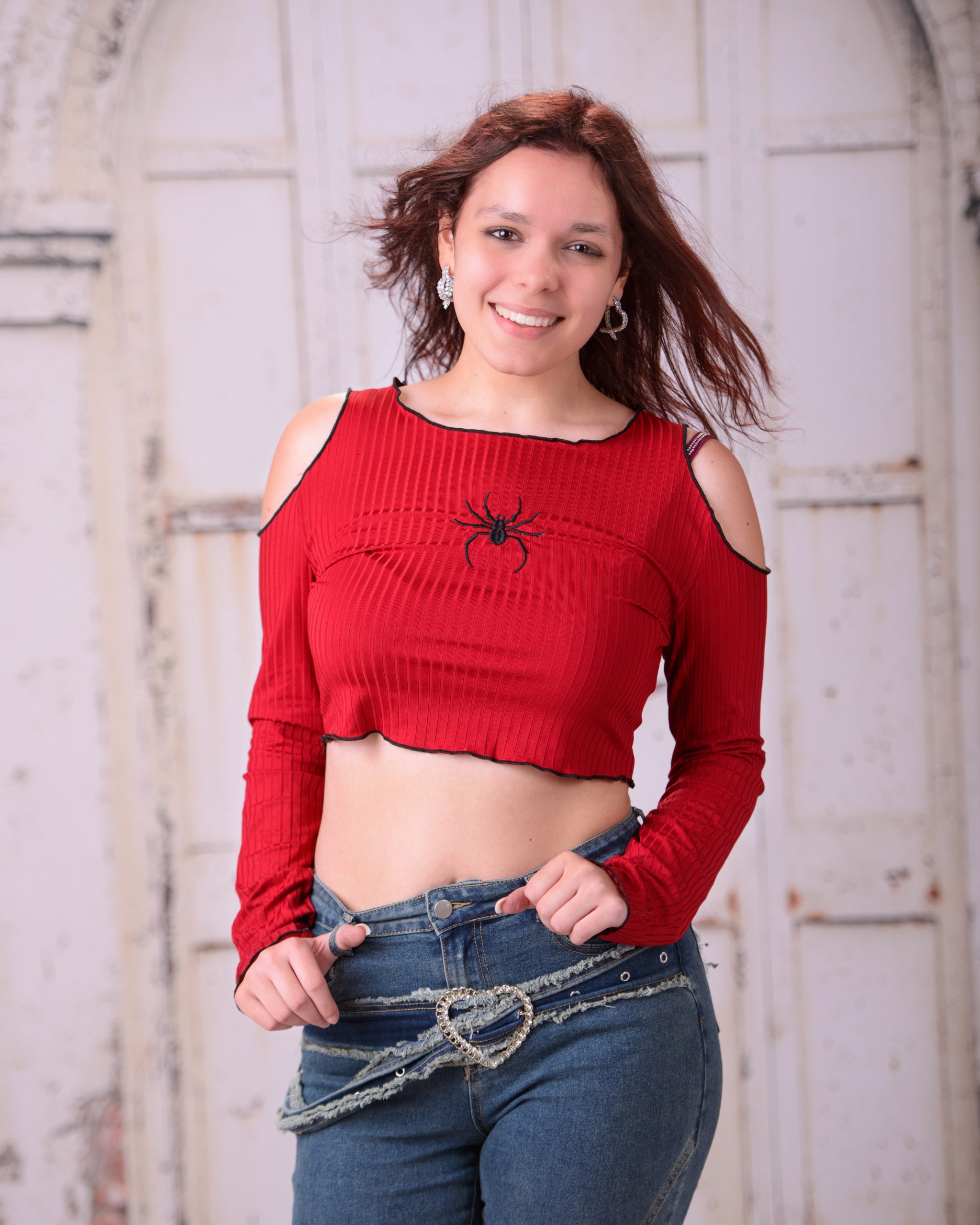 confident senior photo featuring a woman in red off-shoulder top and heart belt detail standing before weathered white doors