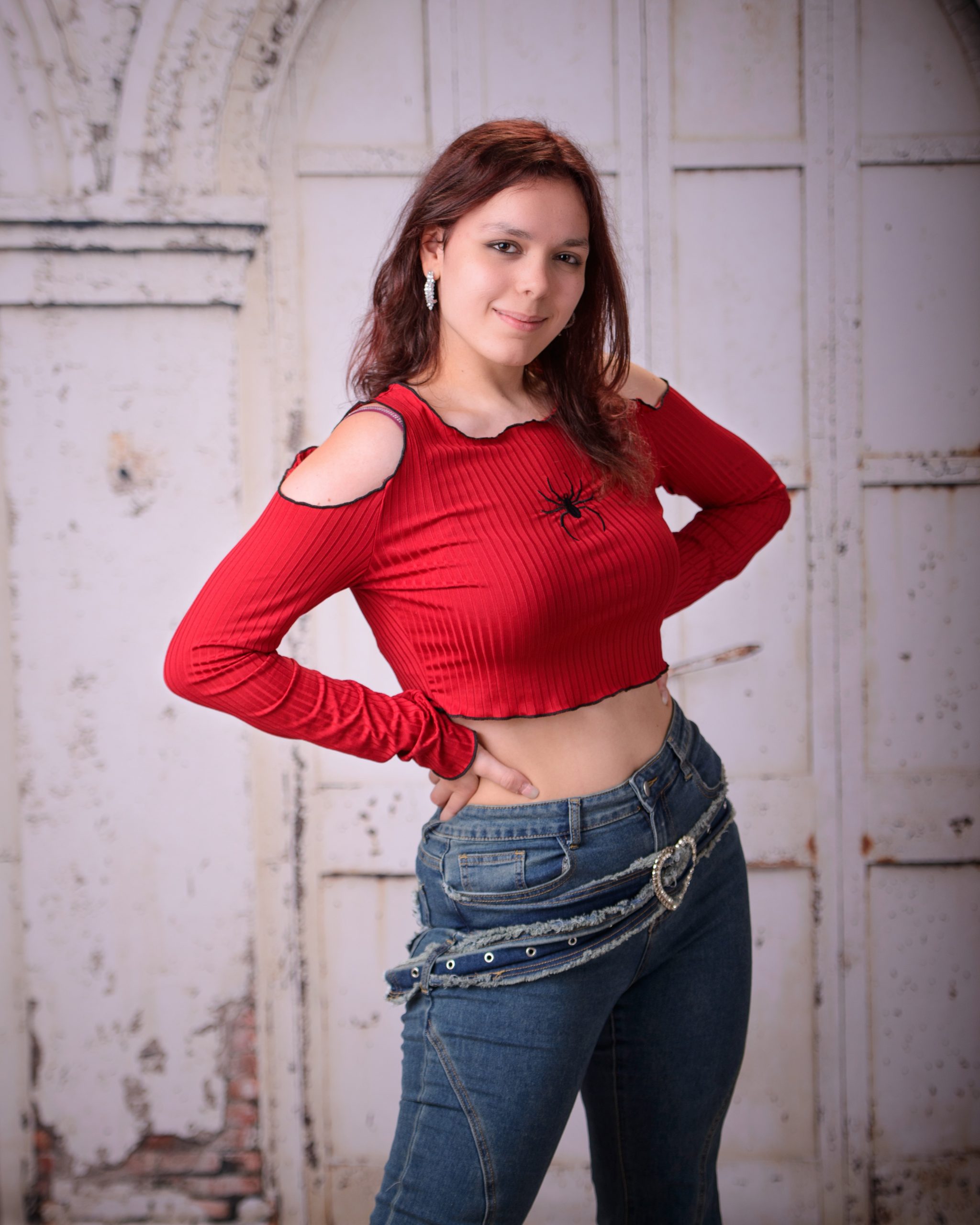 confident senior photo featuring a woman in red off-shoulder top and heart belt detail standing before weathered white doors