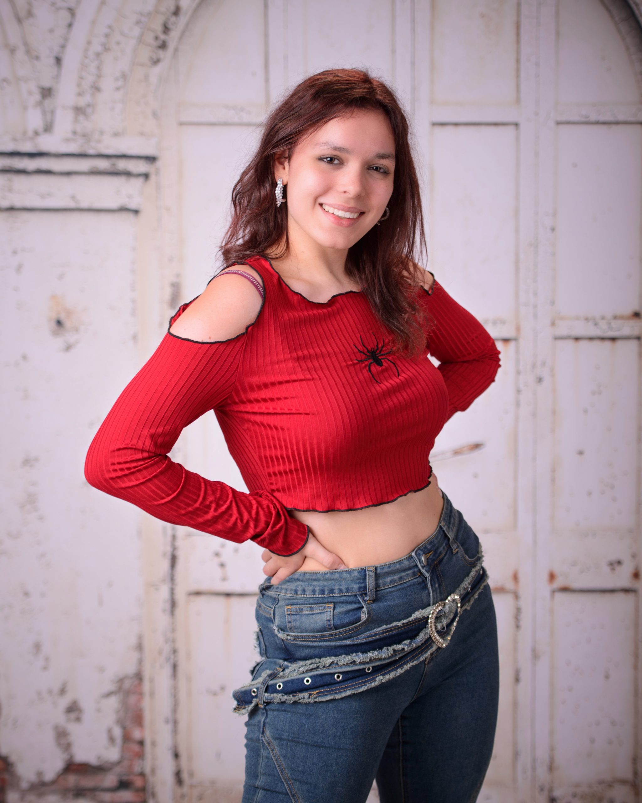 confident senior photo featuring a woman in red off-shoulder top and heart belt detail standing before weathered white doors