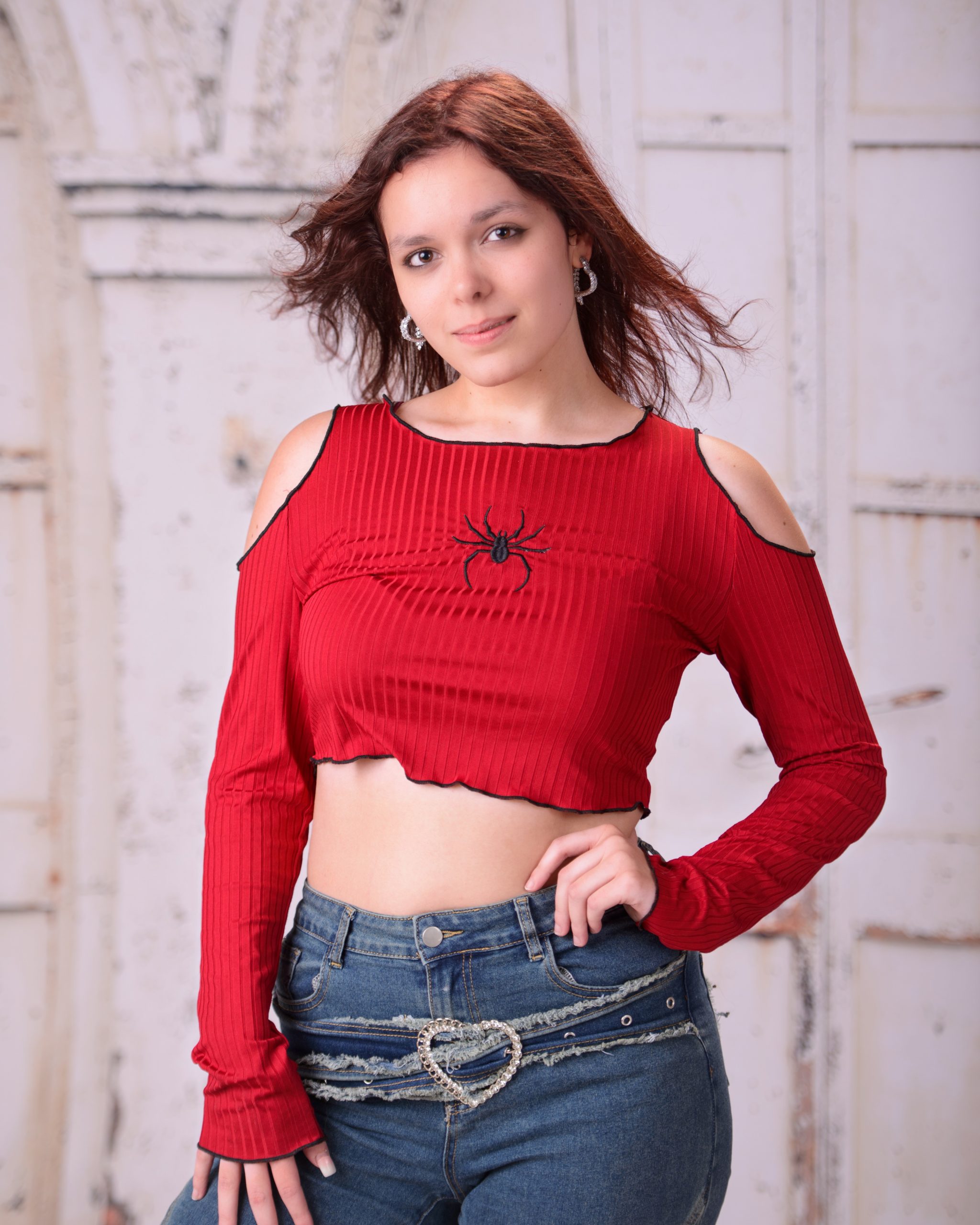 confident senior photo featuring a woman in red off-shoulder top and heart belt detail standing before weathered white doors