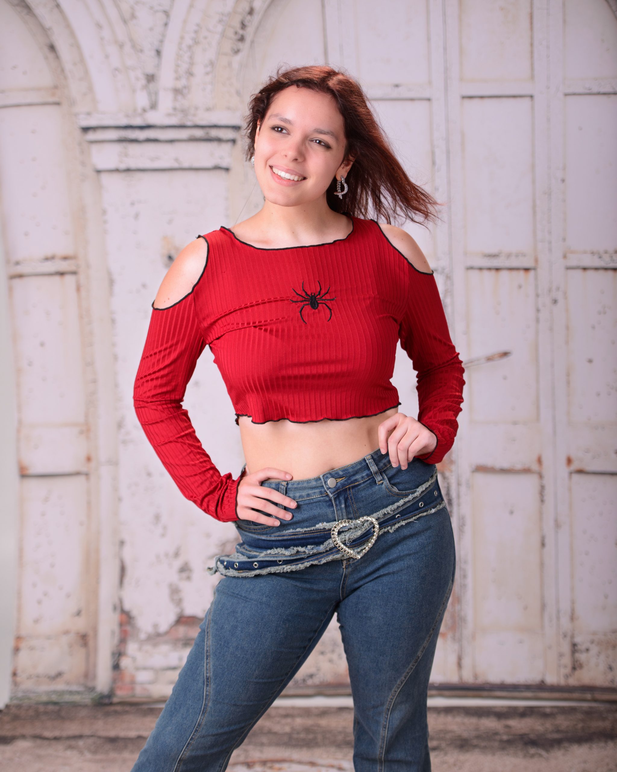 confident senior photo featuring a woman in red off-shoulder top and heart belt detail standing before weathered white doors