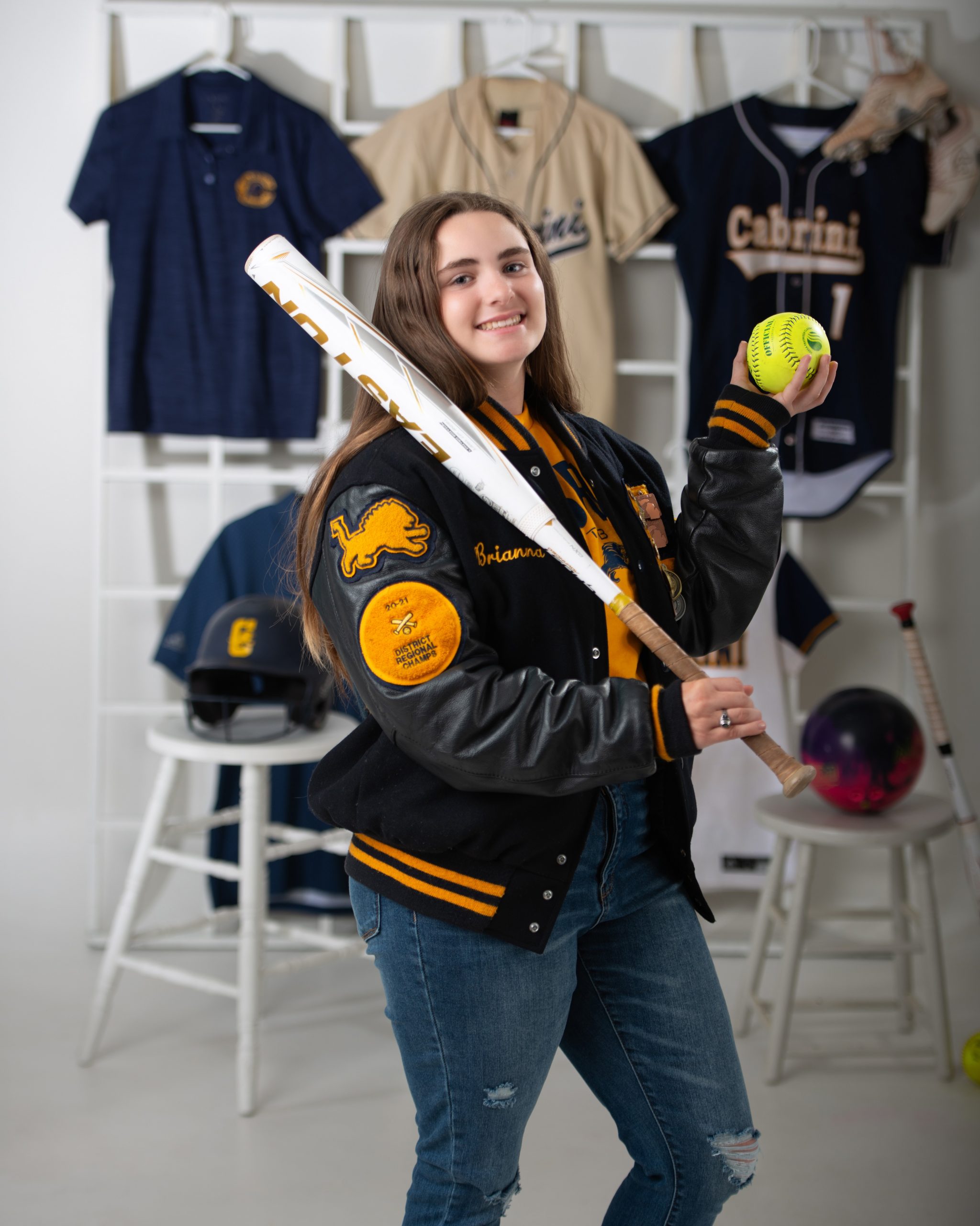 Branna wearing graduation cap and gown holding softball bat and ball, sports jerseys in background for senior sports photography
