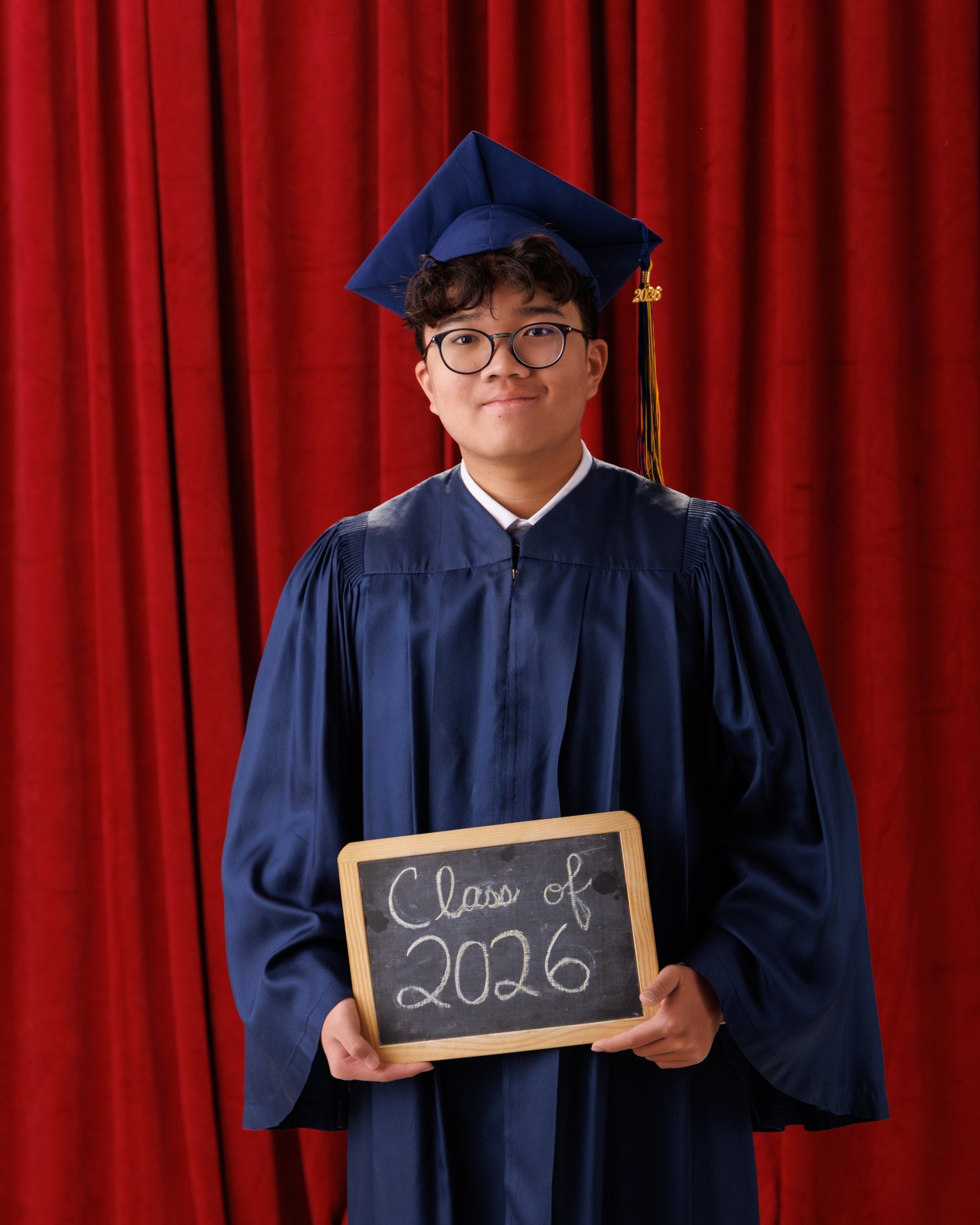 Graduating senior in blue gown holding 'Class of 2026' chalkboard sign, wearing cap with tassel, red curtain backdrop