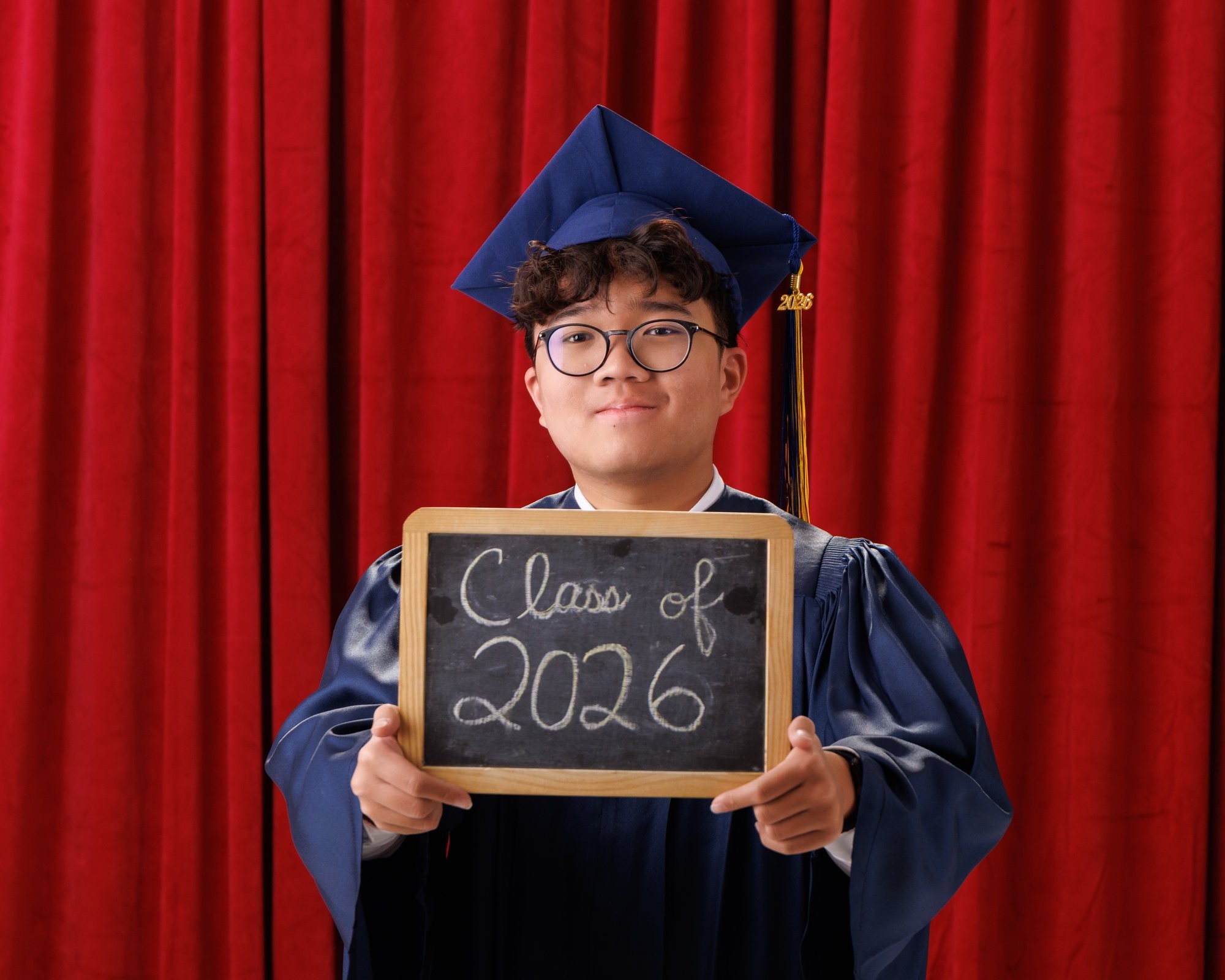 Senior in graduation attire holding 'Class of 2026' chalkboard, navy gown and cap, red curtains