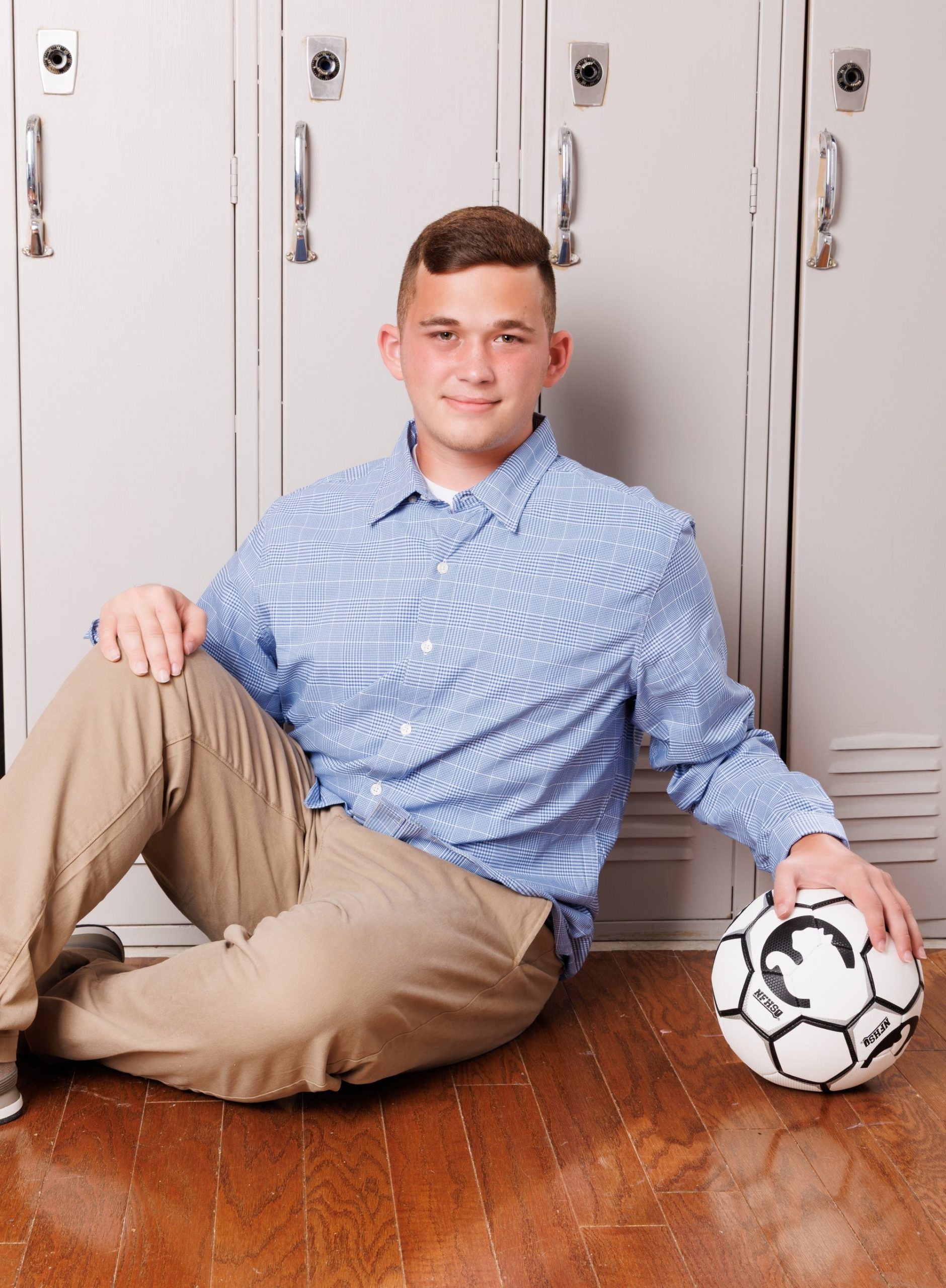 Senior Joseph holds soccer ball while seated on wooden floor with lockers background