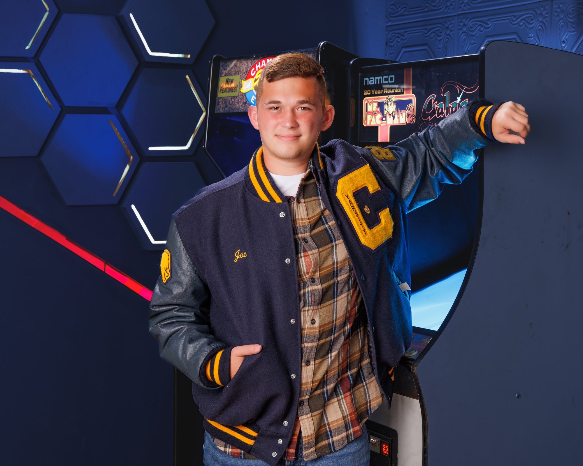 Senior Joseph in varsity jacket stands before arcade machines, featuring 'Cabrini' branding