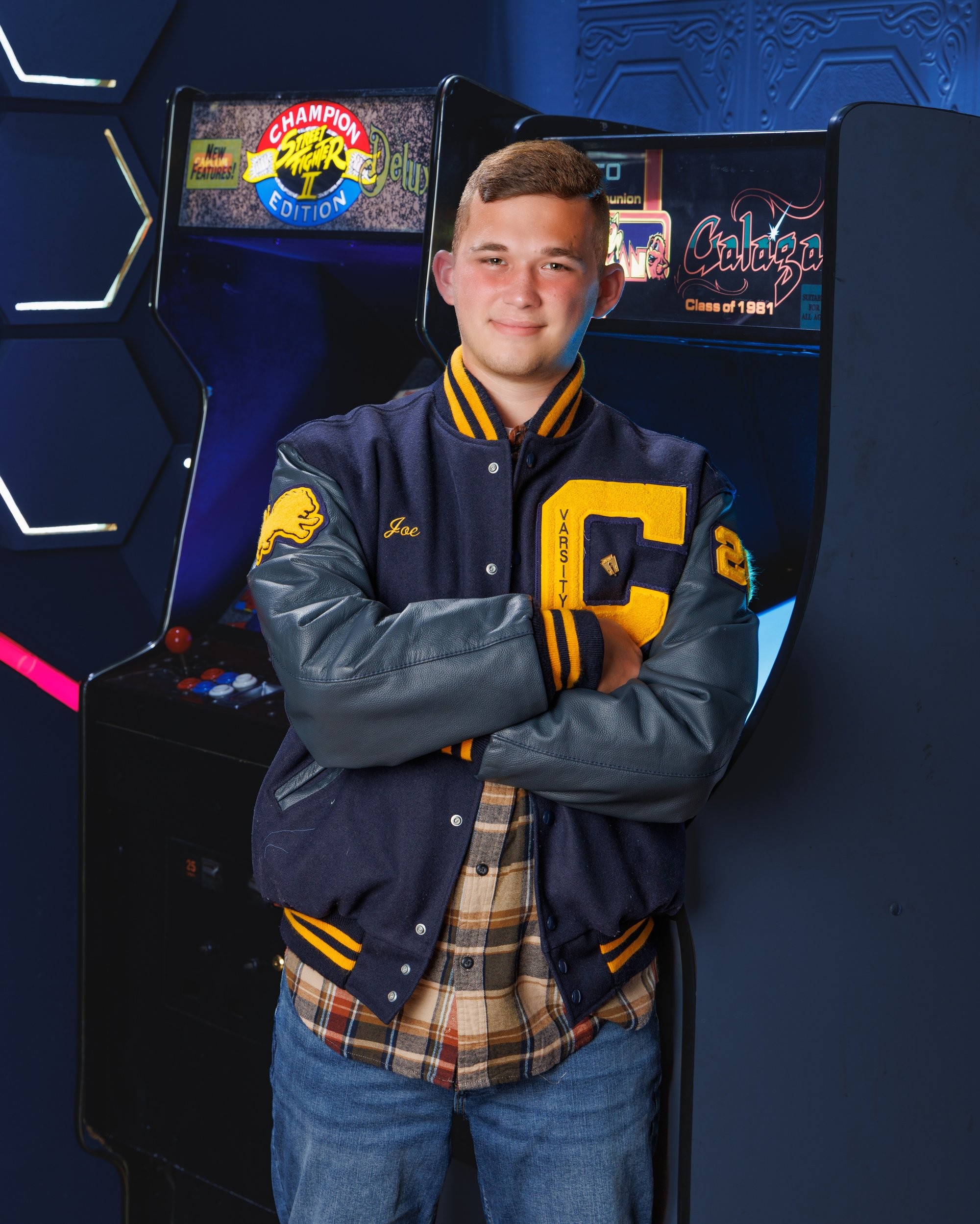 Senior Joseph in varsity jacket stands before arcade machines, featuring 'Cabrini' branding