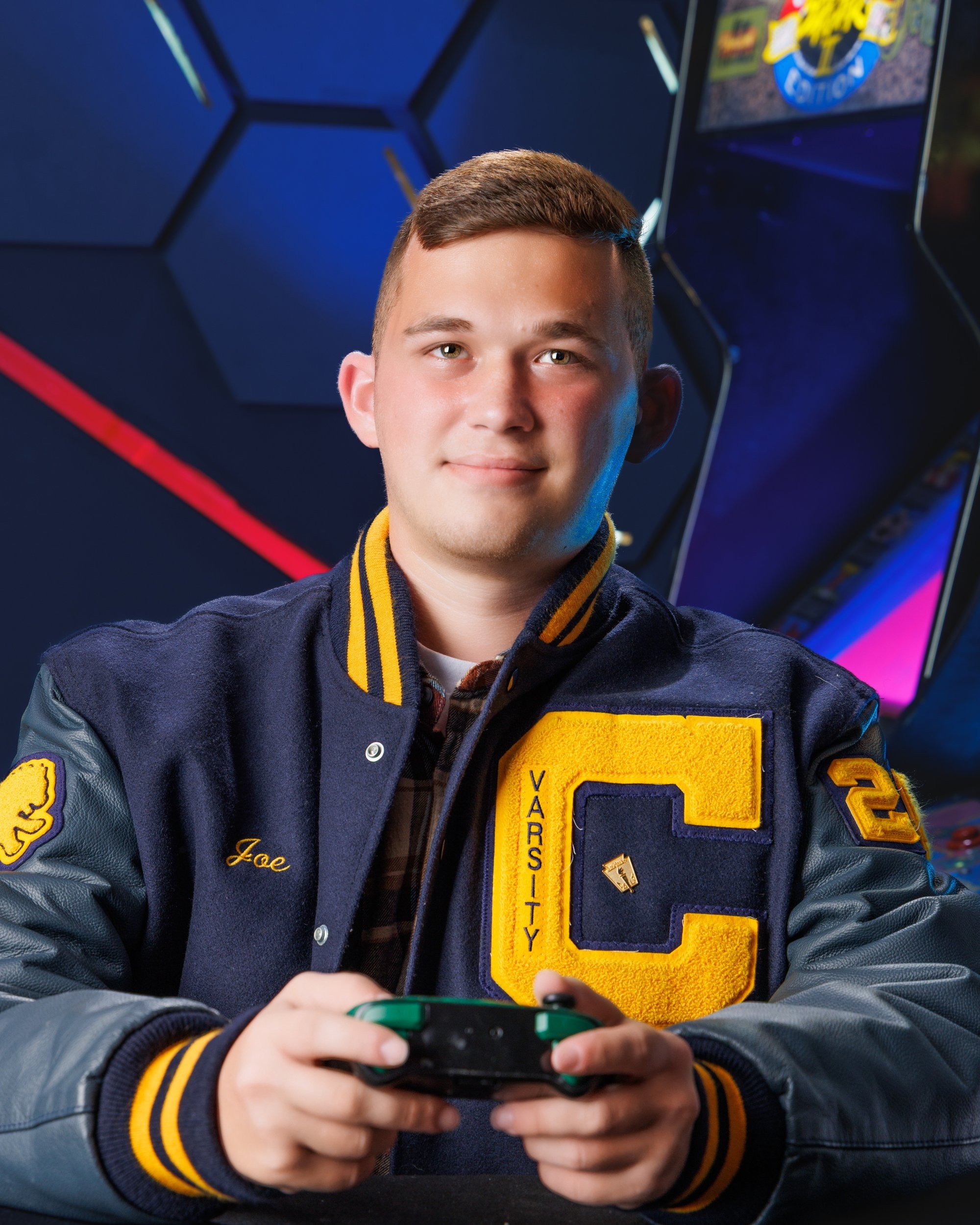 Senior Joseph in varsity jacket stands before arcade machines, featuring 'Cabrini' branding