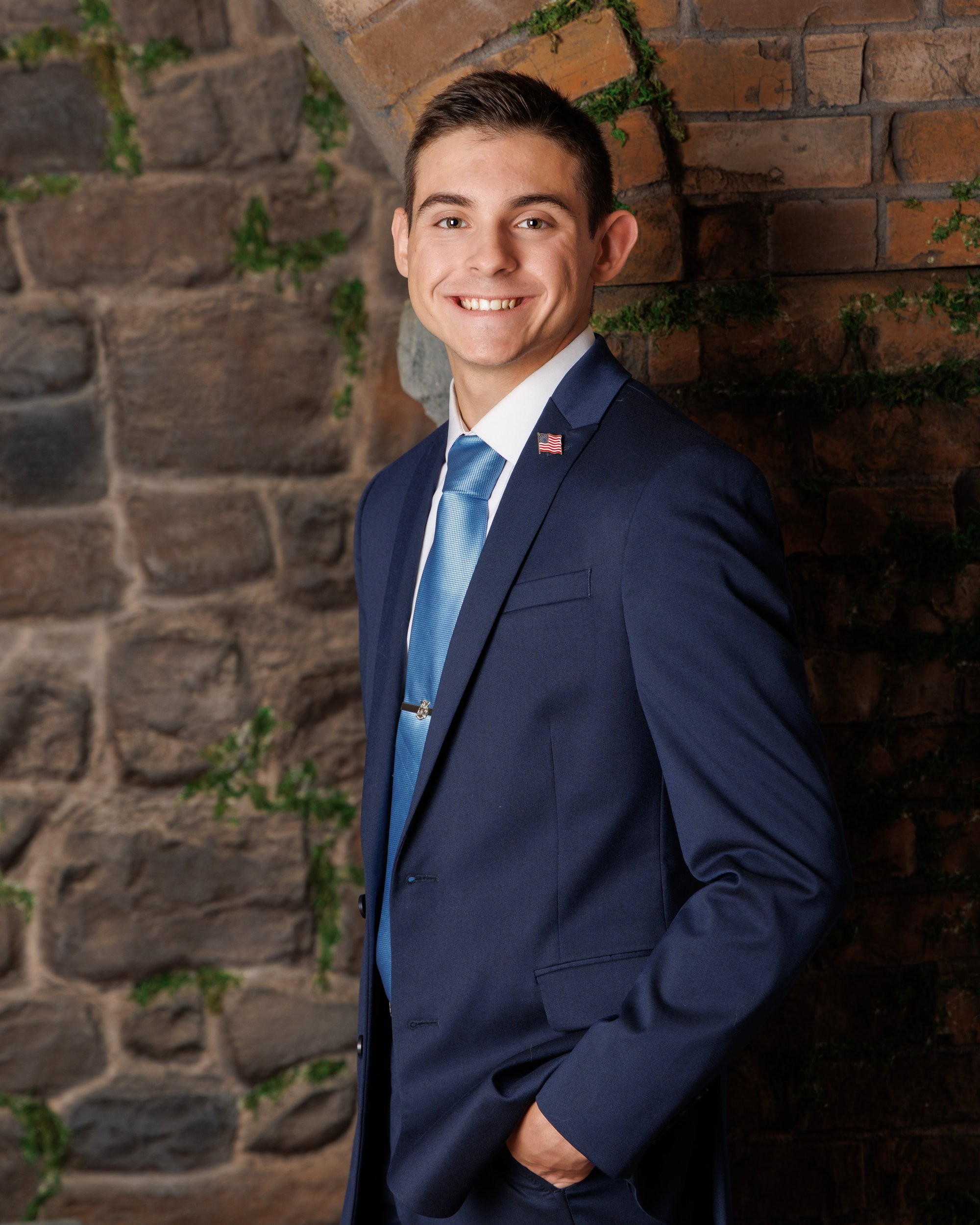 Young man in black zip-up jacket and green tie seated on stone ledge, background features brick and ivy.