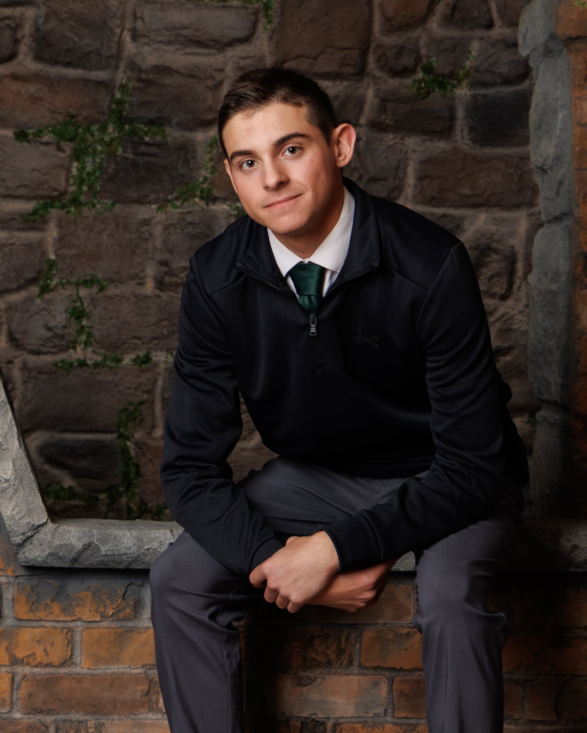 Senior portrait of a young adult in black zip-up and green tie standing against textured stone wall.