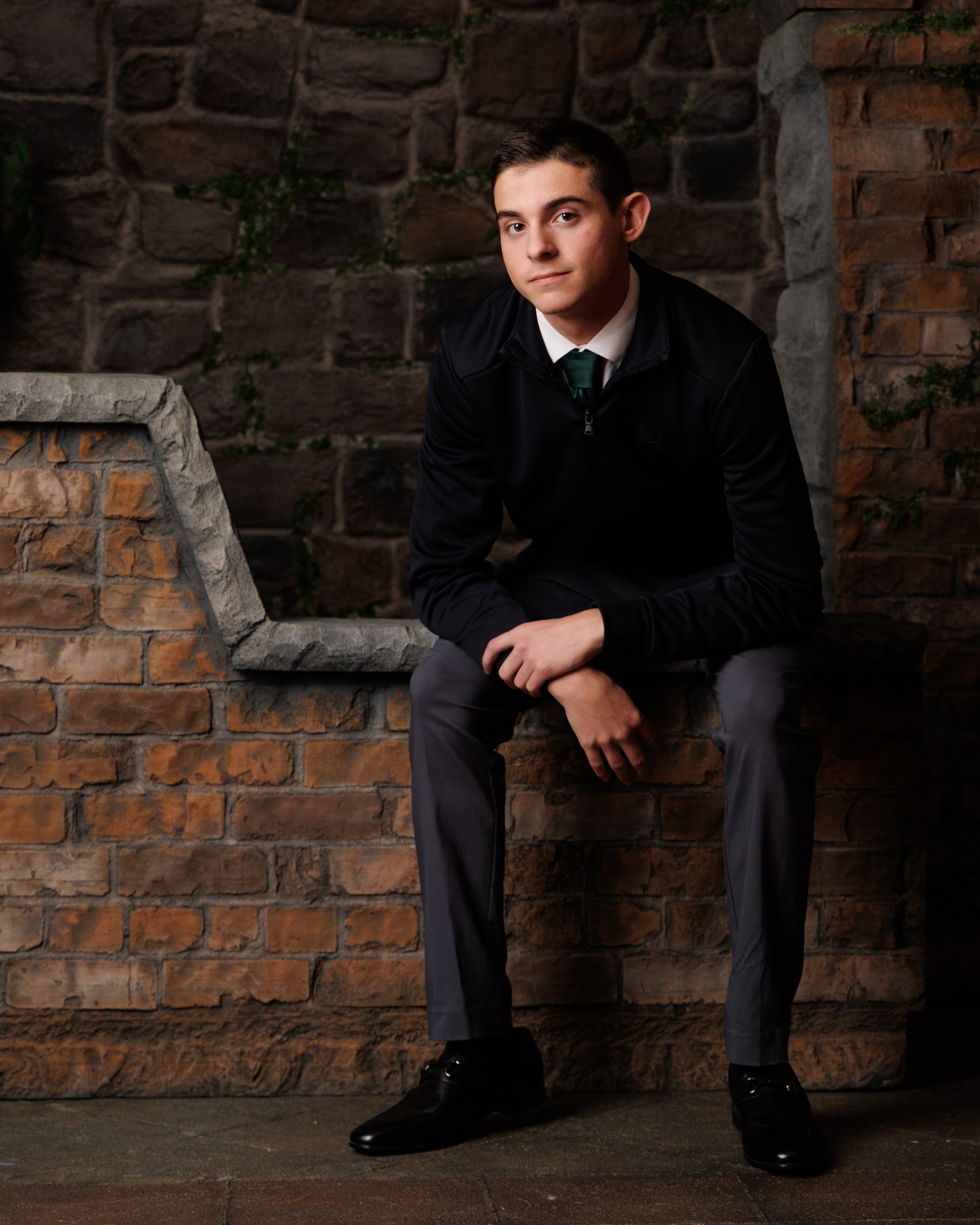 Senior portrait of a young adult in black zip-up and green tie standing against textured stone wall.