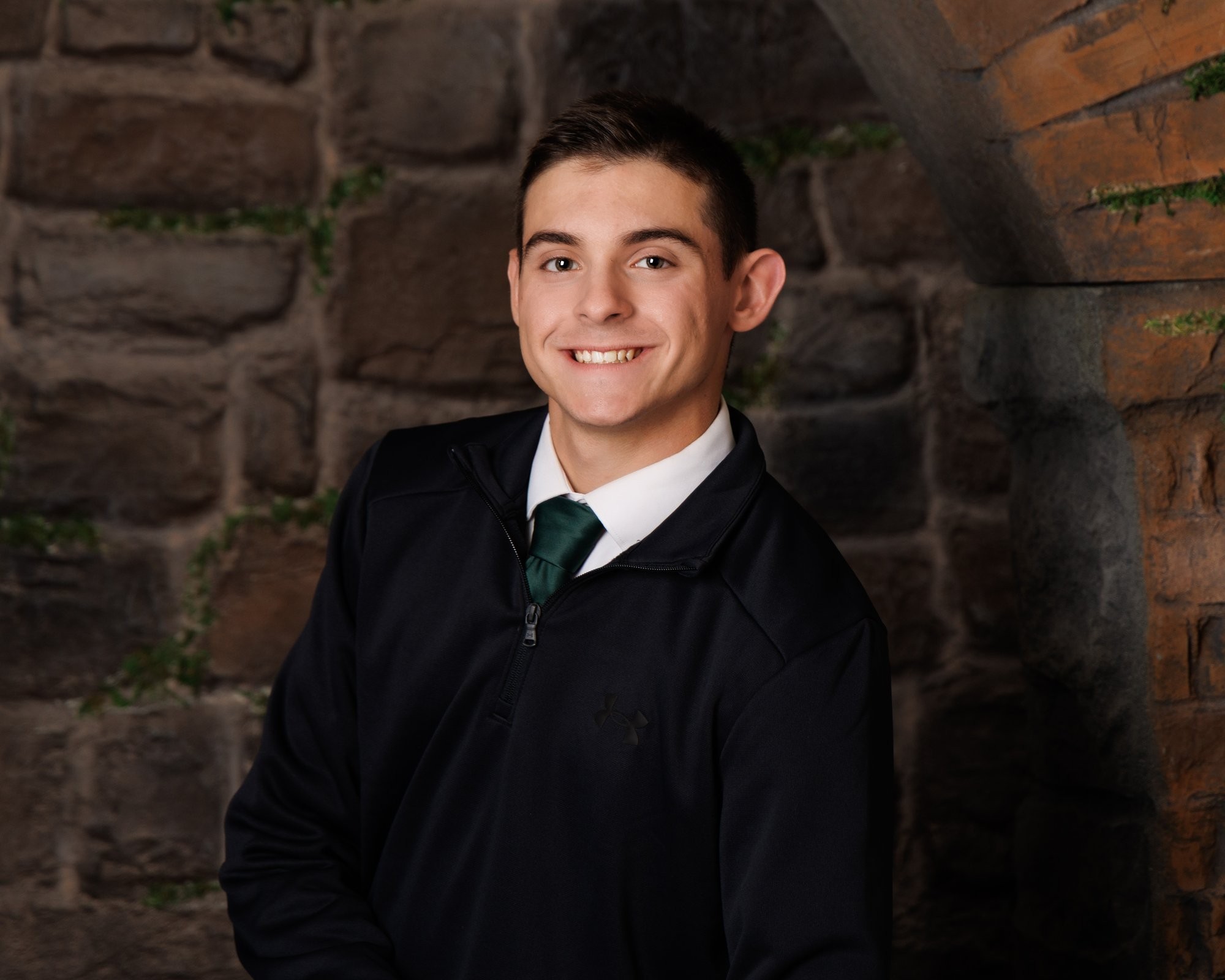 Senior portrait of a young adult in black zip-up and green tie standing against textured stone wall.