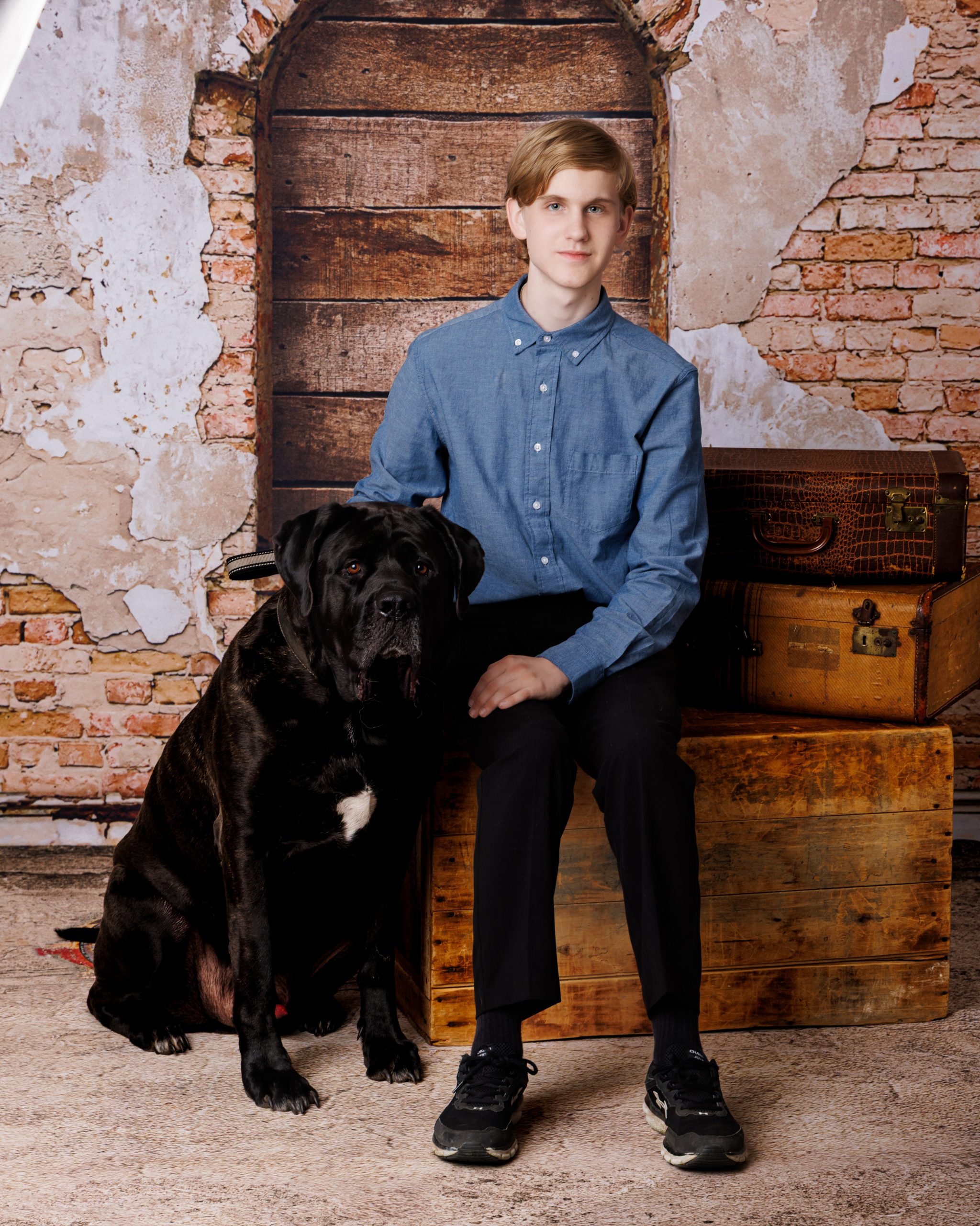 A young man sits beside a large black dog on vintage luggage in front of a rustic brick wall backdrop