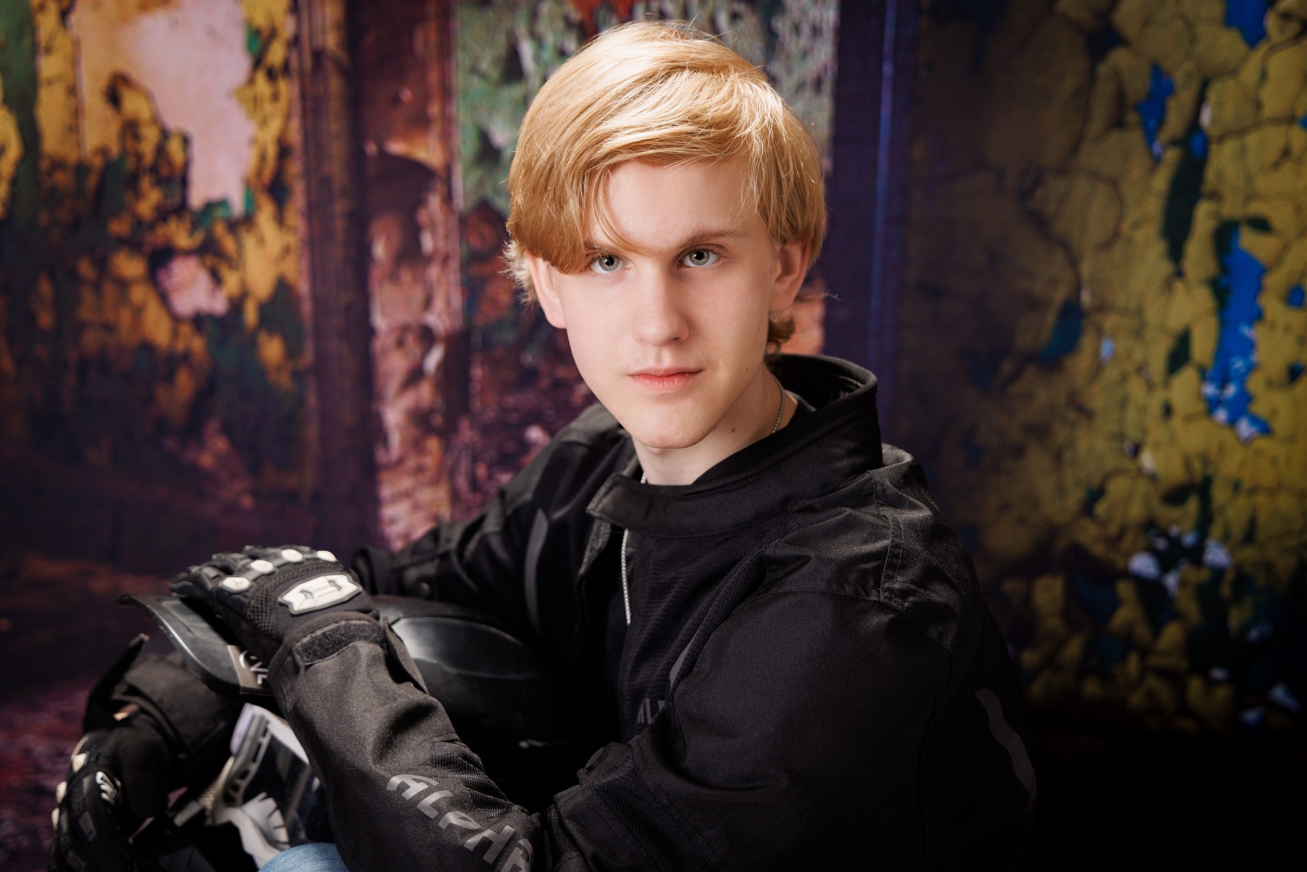 A senior holds a motorcross helmet and goggles while standing against a vibrant, peeling wall backdrop