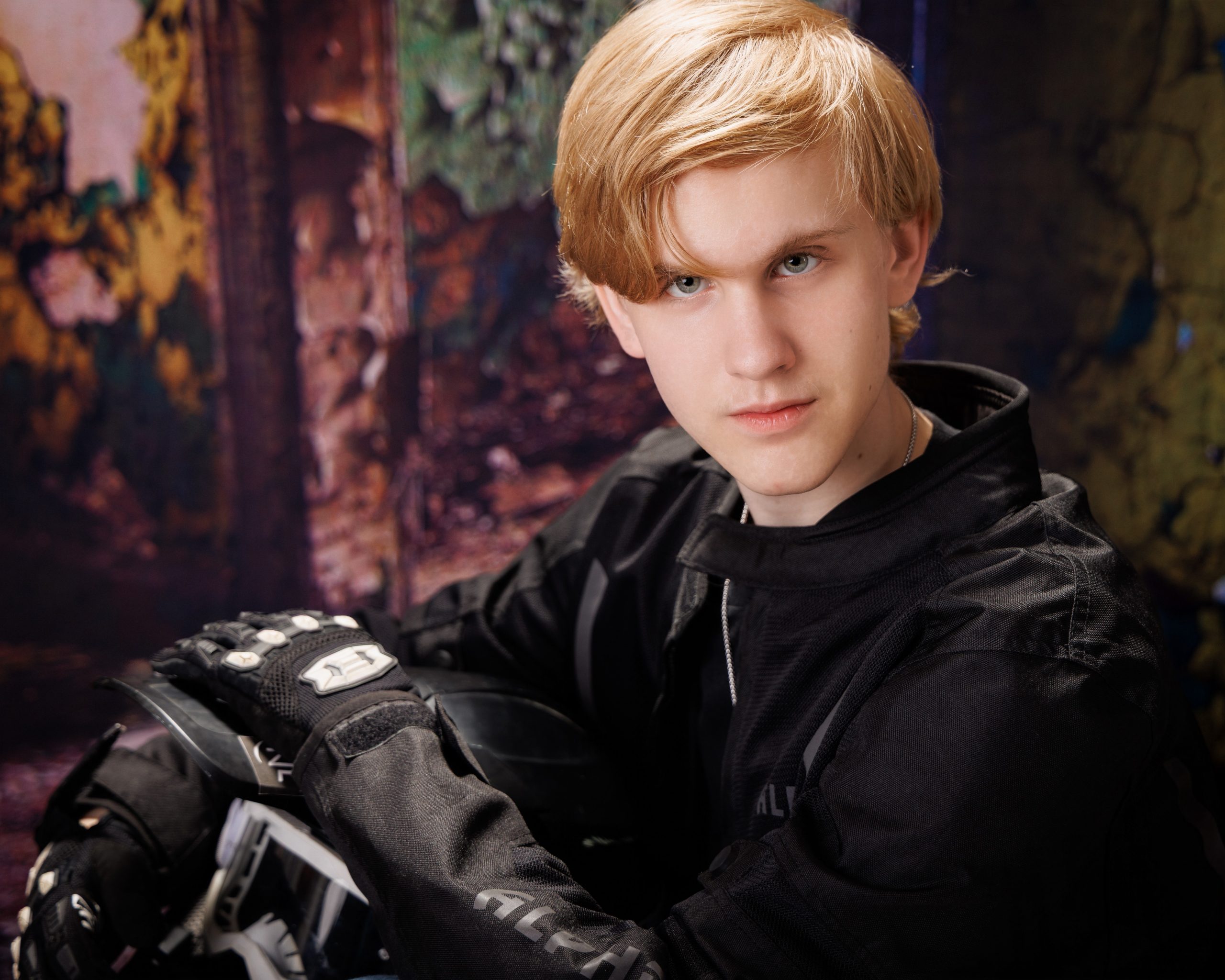 A senior holds a motorcross helmet and goggles while standing against a vibrant, peeling wall backdrop