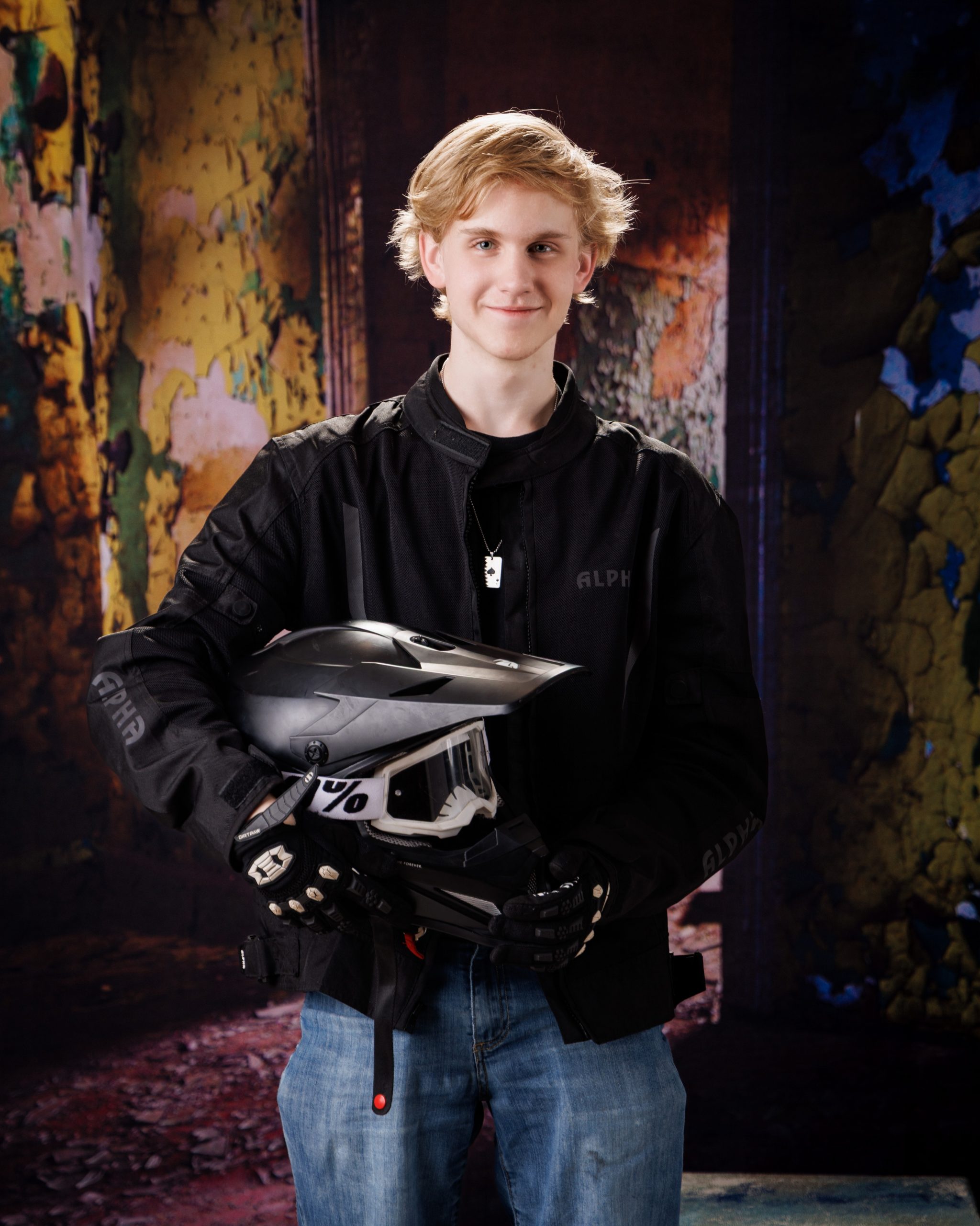 A senior holds a motorcross helmet and goggles while standing against a vibrant, peeling wall backdrop
