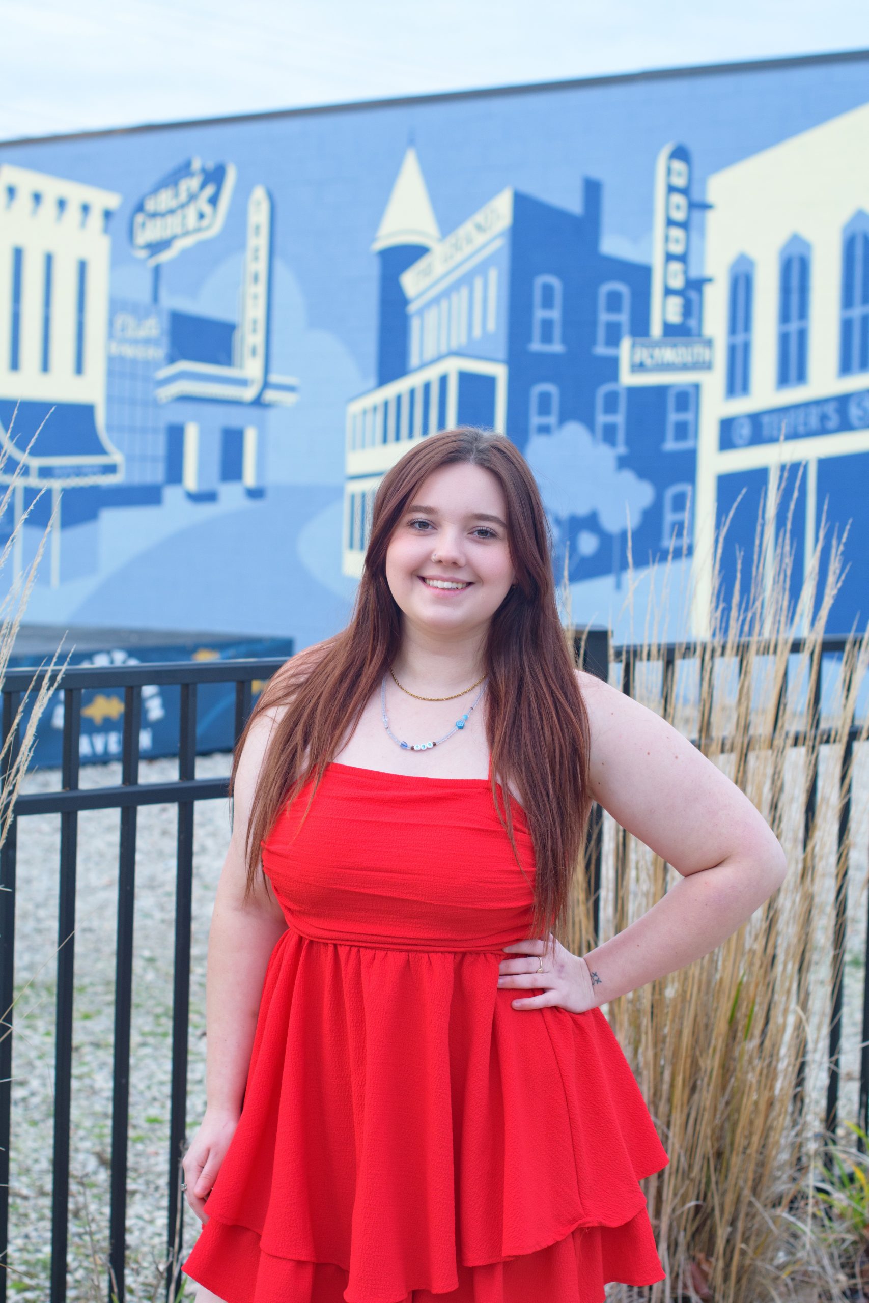 High school student smiling in red dress against a blue cityscape backdrop for Downriver senior photography