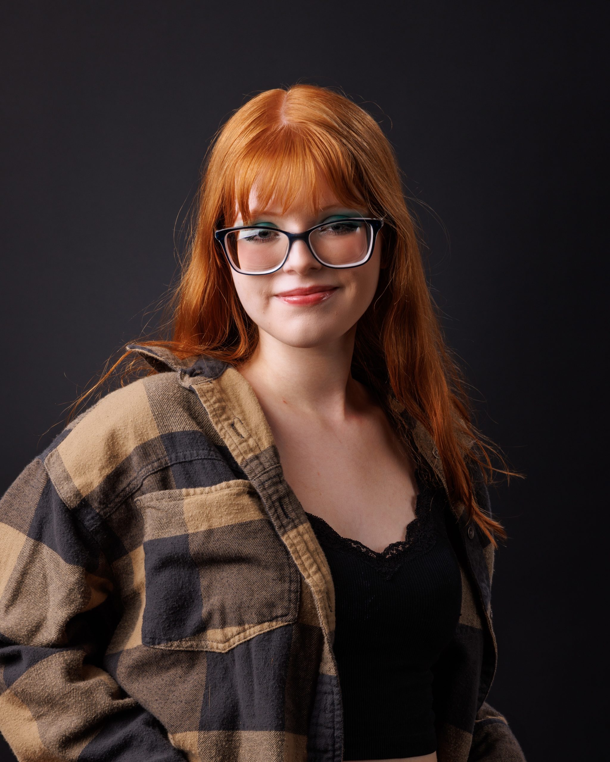 Red-haired senior model wearing plaid shirt and glasses against dark background for studio portrait