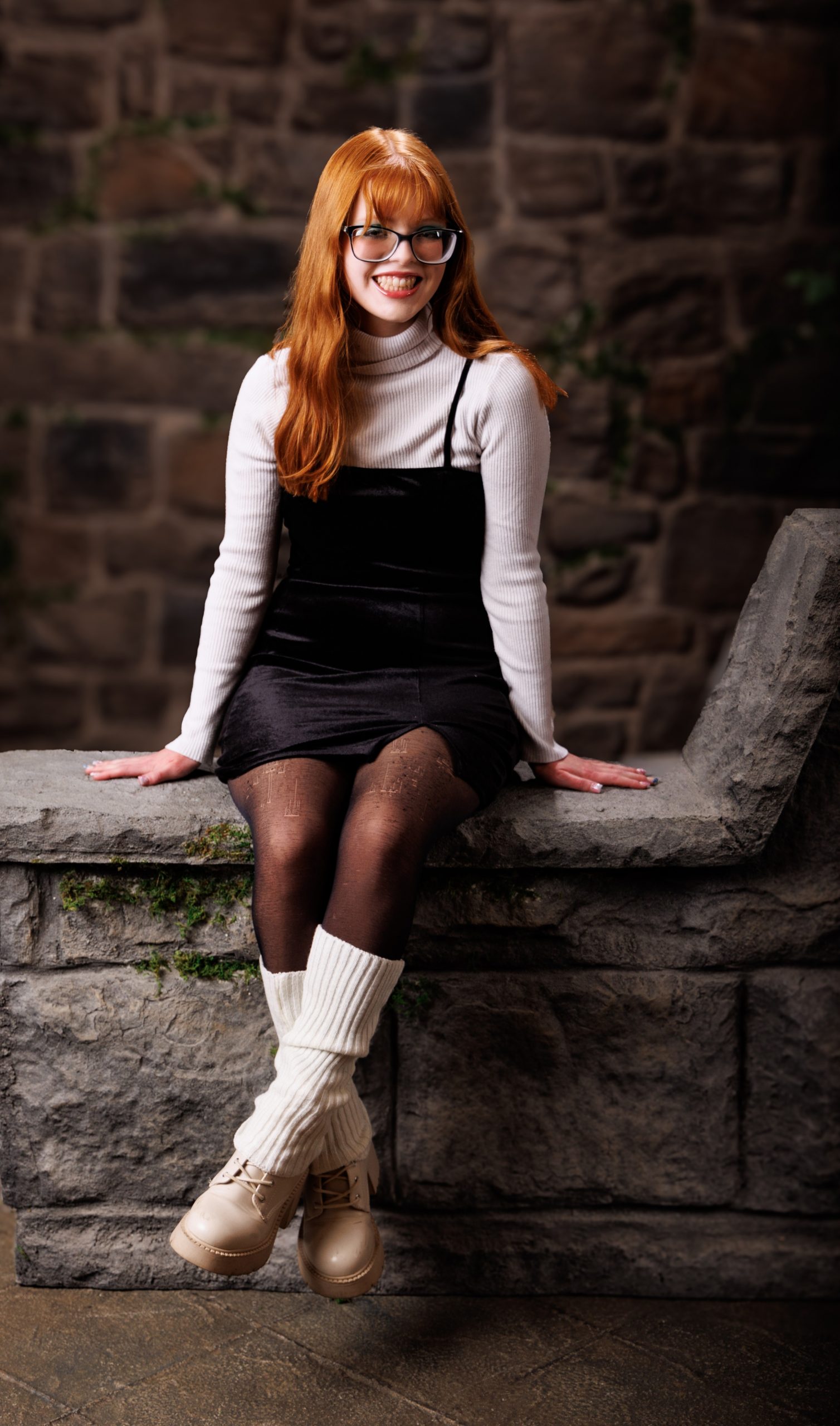 Red-haired model in black velvet dress and turtleneck leaning against stone wall during Wyandotte photo session