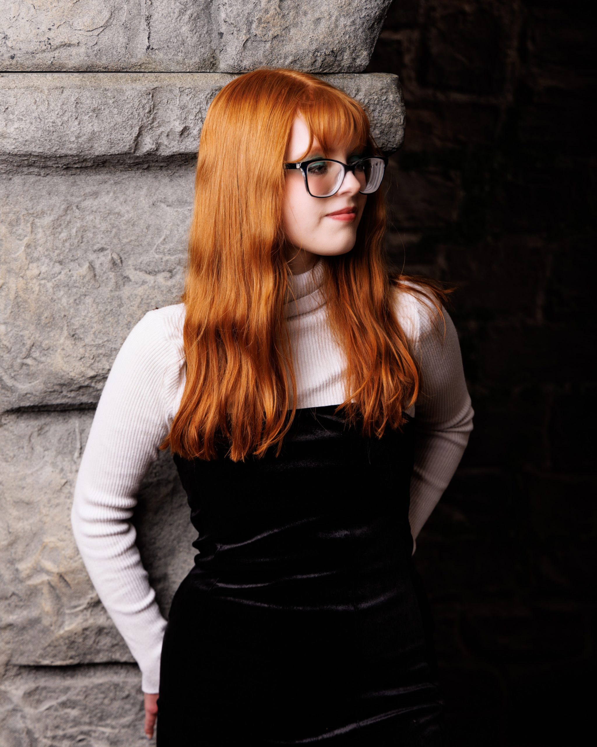 Literary-themed senior portrait of red-haired model wearing glasses and turtleneck against textured stone background