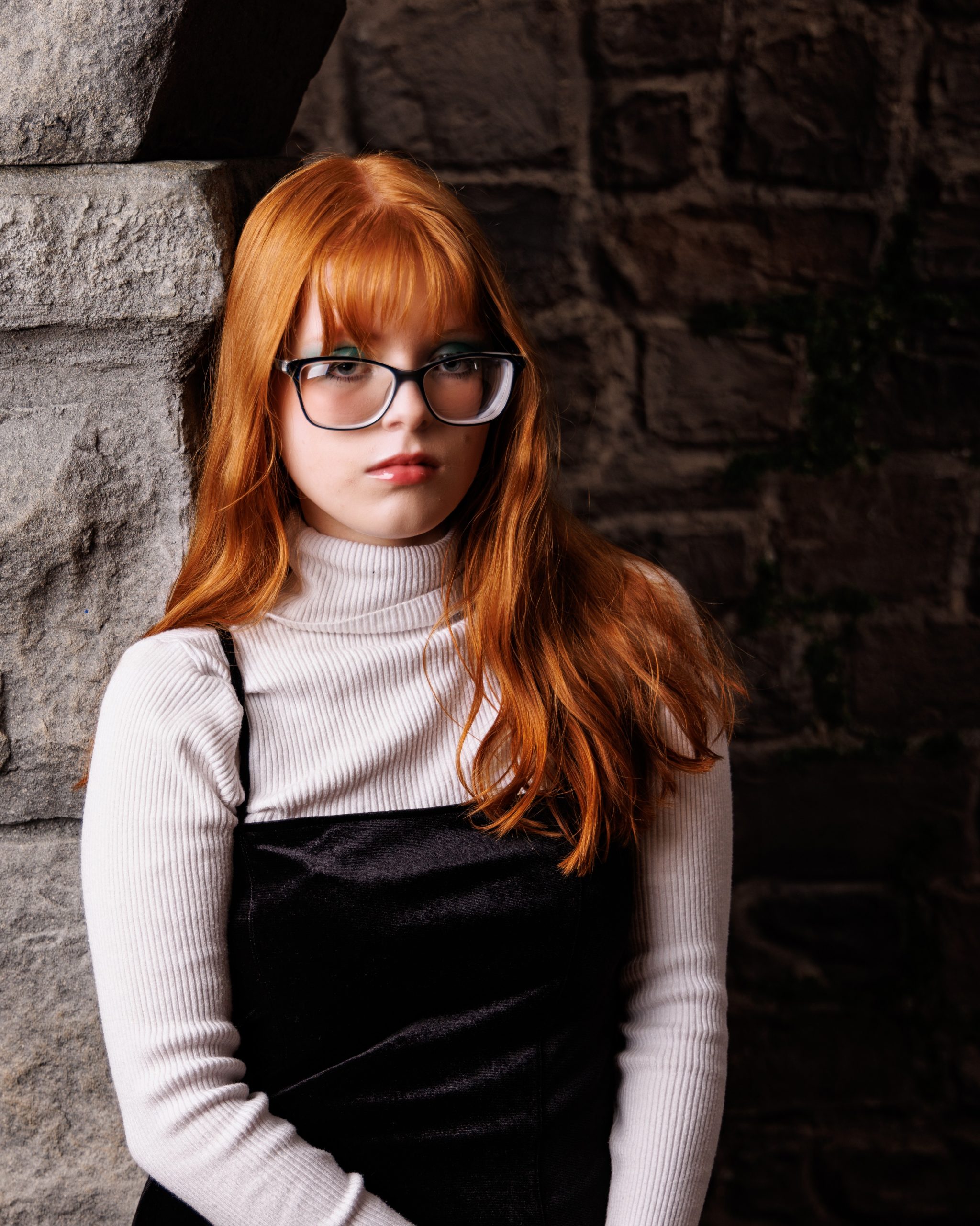 Literary-themed senior portrait of red-haired model wearing glasses and turtleneck against textured stone background
