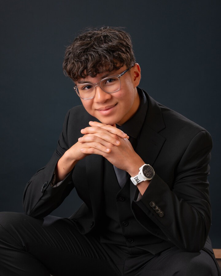 Professional headshot of a young man in a black suit with hands clasped, wearing glasses and a smartwatch, Downriver senior portrait session