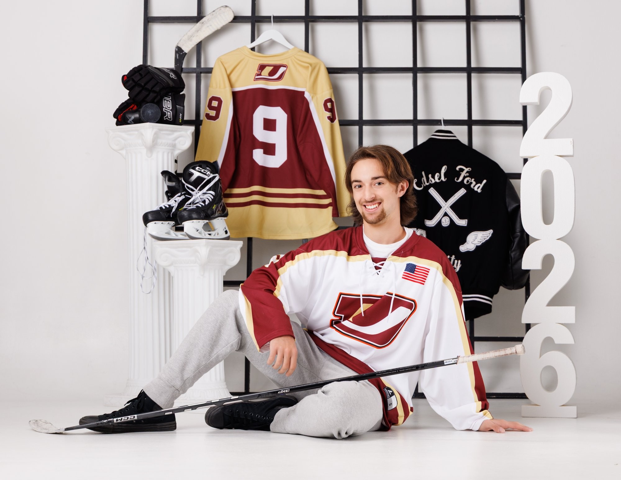 Henry posing with hockey gloves on shoulders and a hockey stick across his back against a chain-link fence background