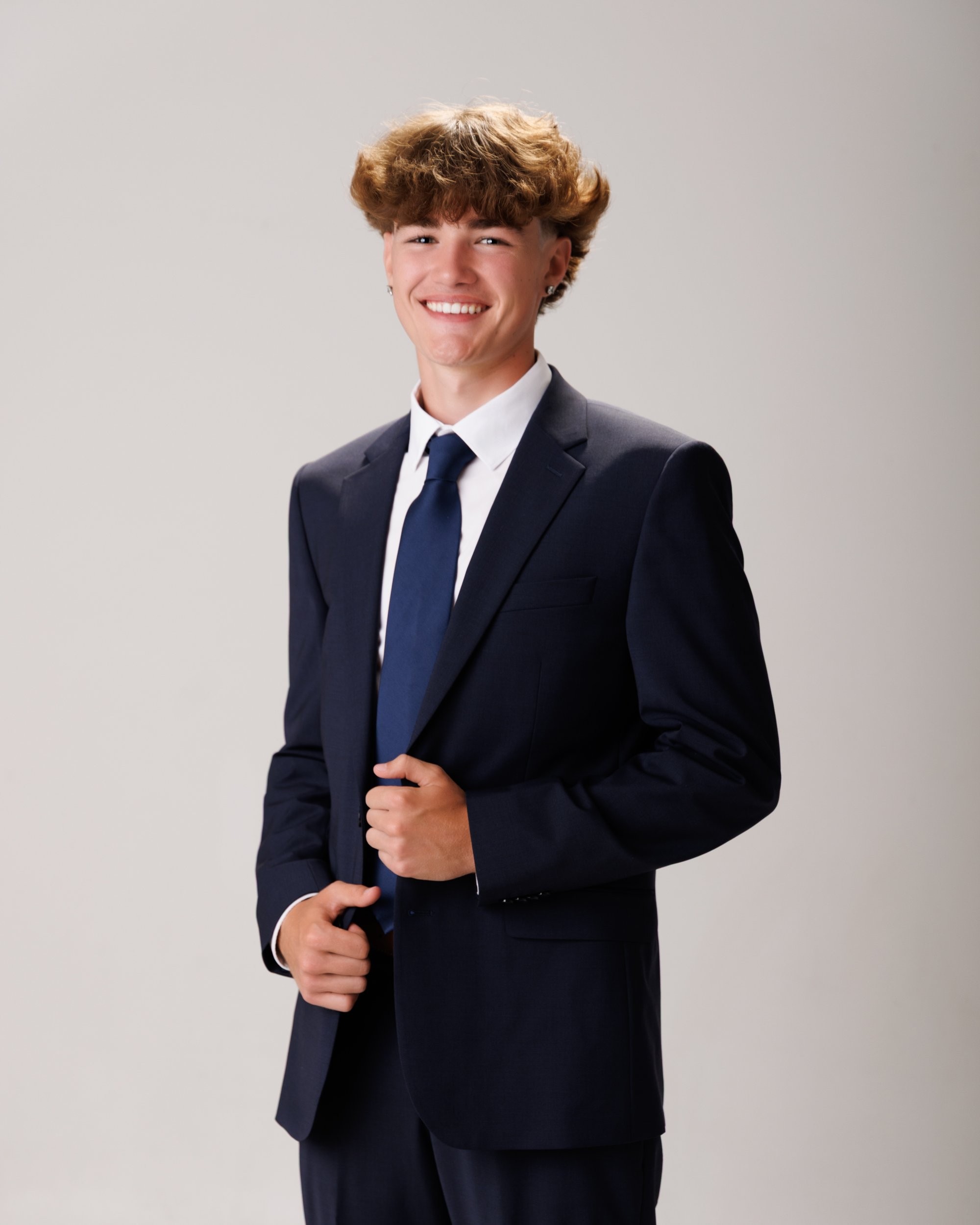 Evan in a navy suit and tie, smiling confidently against a dark studio backdrop for senior photos