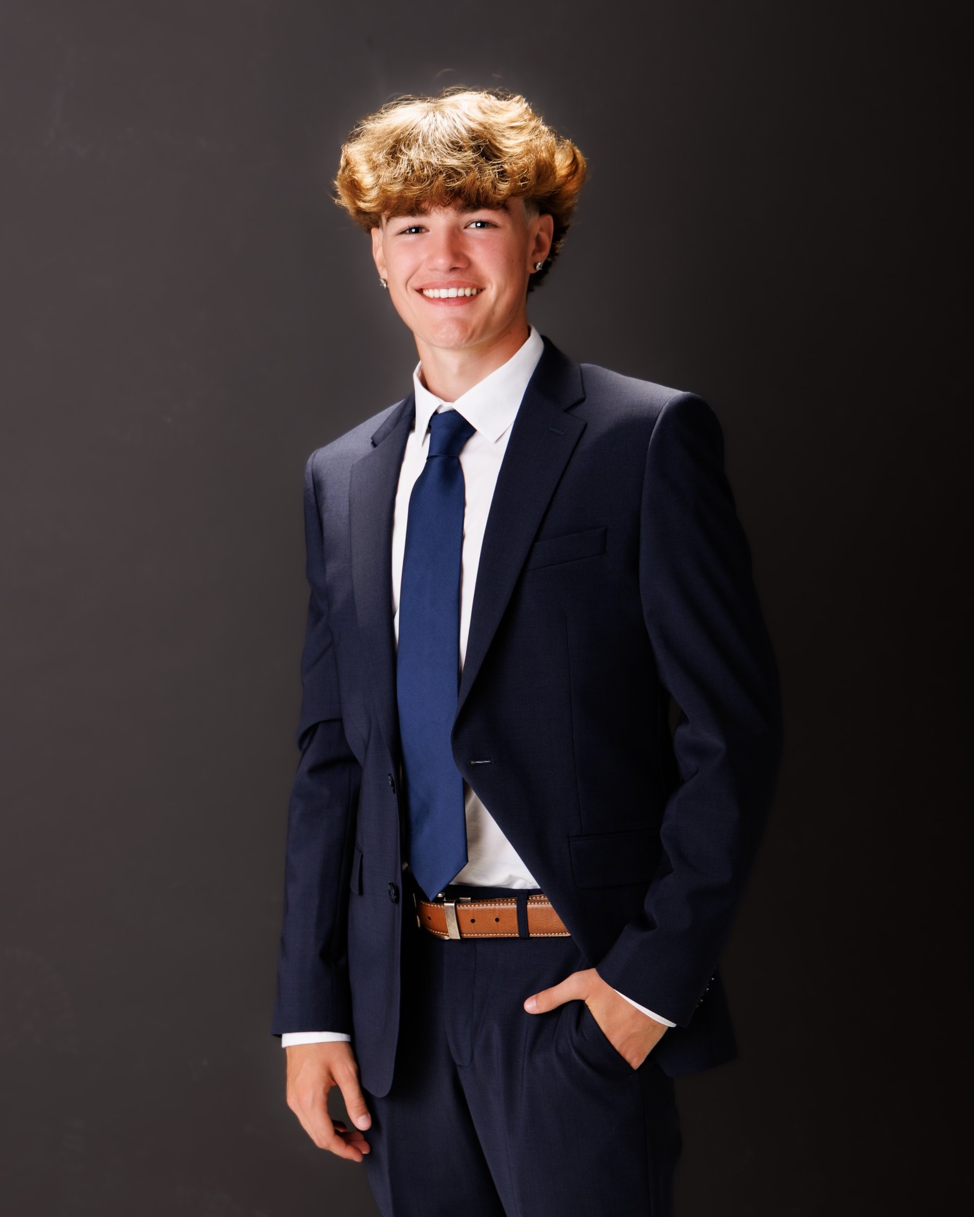 Evan in a navy suit and tie, smiling confidently against a dark studio backdrop for senior photos