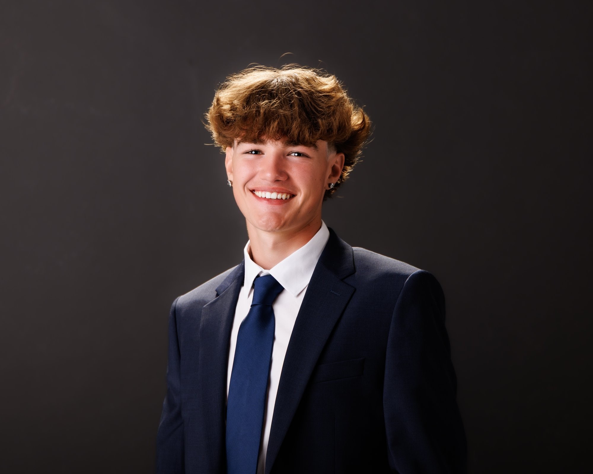 Evan in a navy suit and tie, smiling confidently against a dark studio backdrop for senior photos
