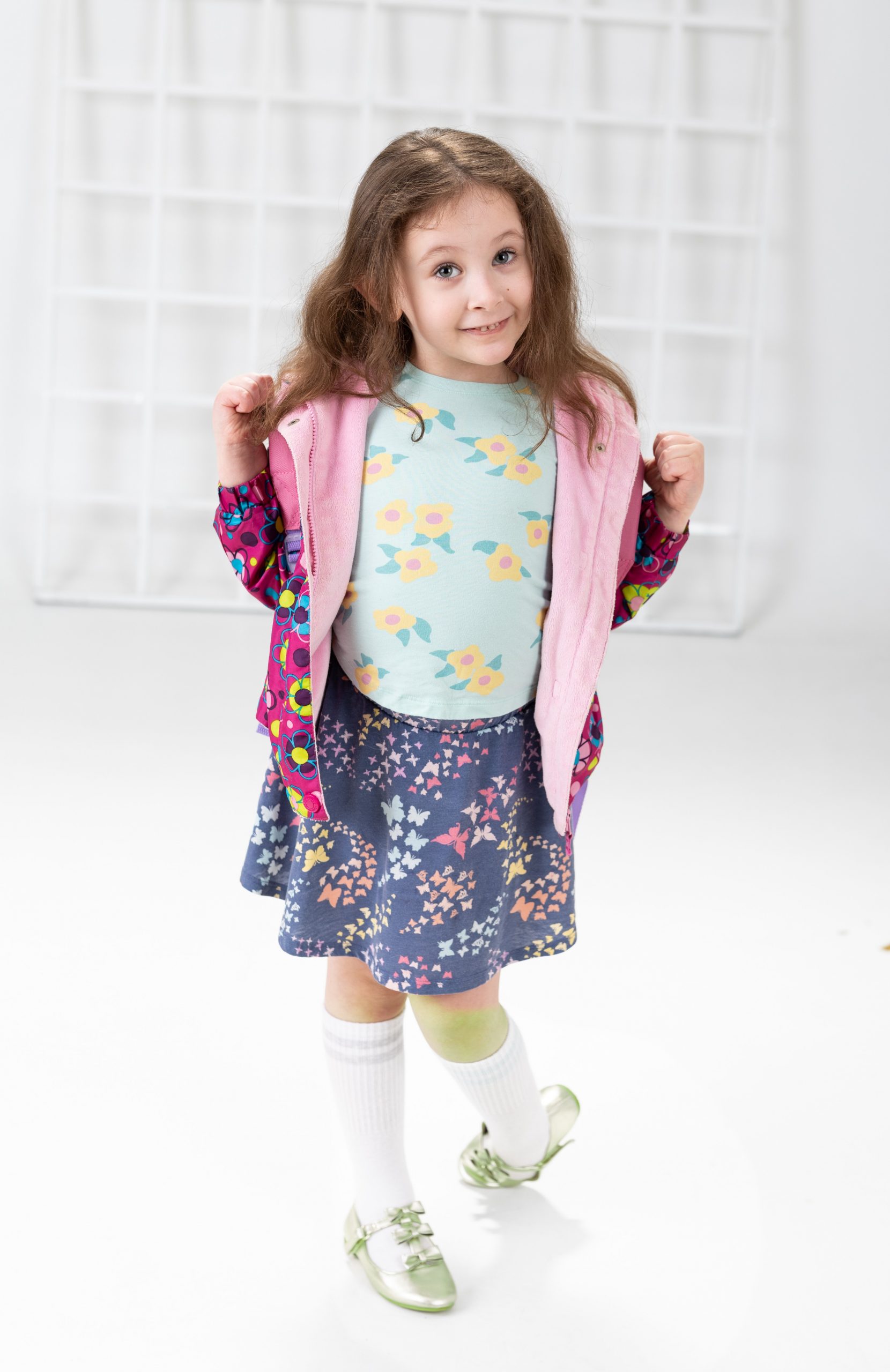 Five-year-old girl wearing light blue floral shirt, navy butterfly skirt, pink jacket, green shoes, holding jacket straps