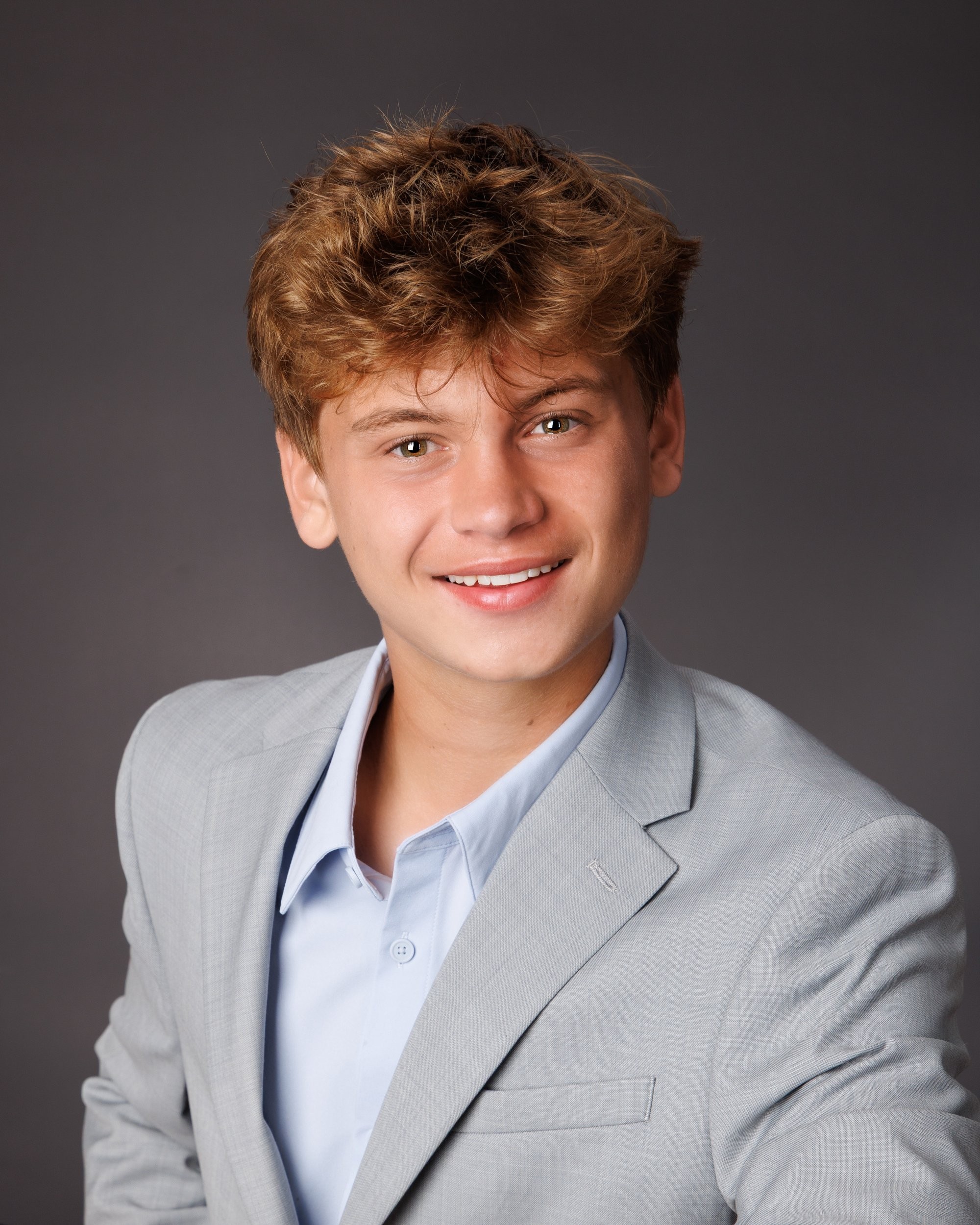 Grosse Isle senior photo showing Luke adjusting his blazer, hands near chest, in light gray suit and blue shirt on dark background