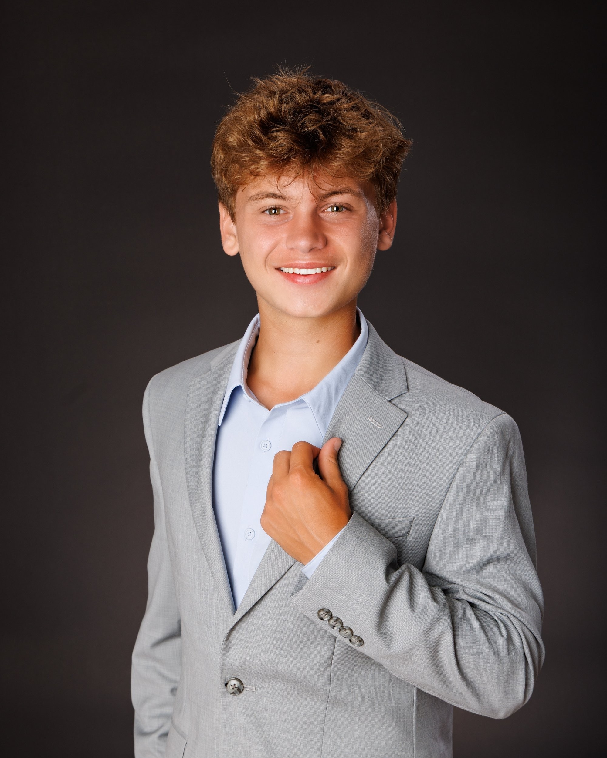 Grosse Isle senior photo showing Luke adjusting his blazer, hands near chest, in light gray suit and blue shirt on dark background