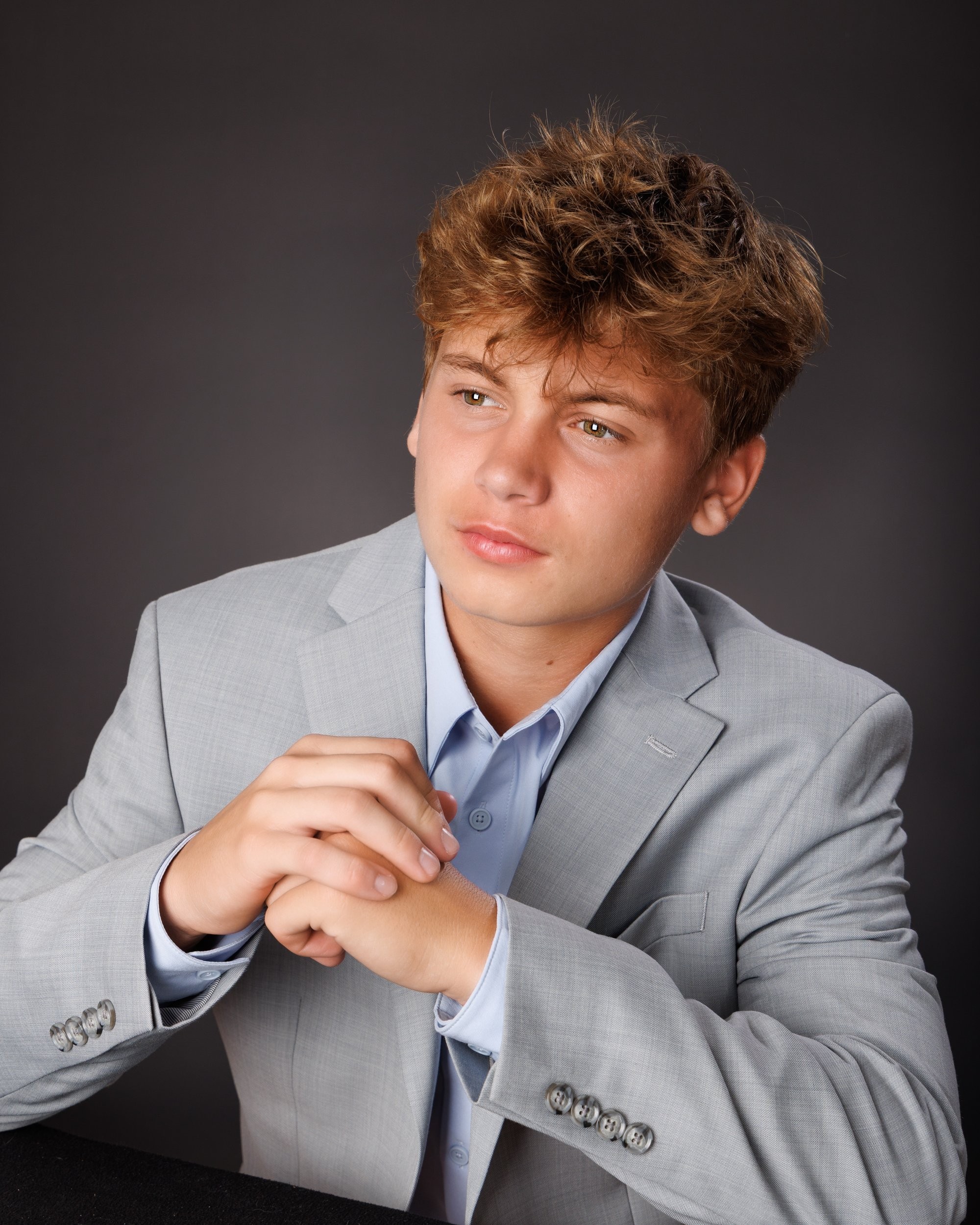 Grosse Isle senior photo showing Luke adjusting his blazer, hands near chest, in light gray suit and blue shirt on dark background