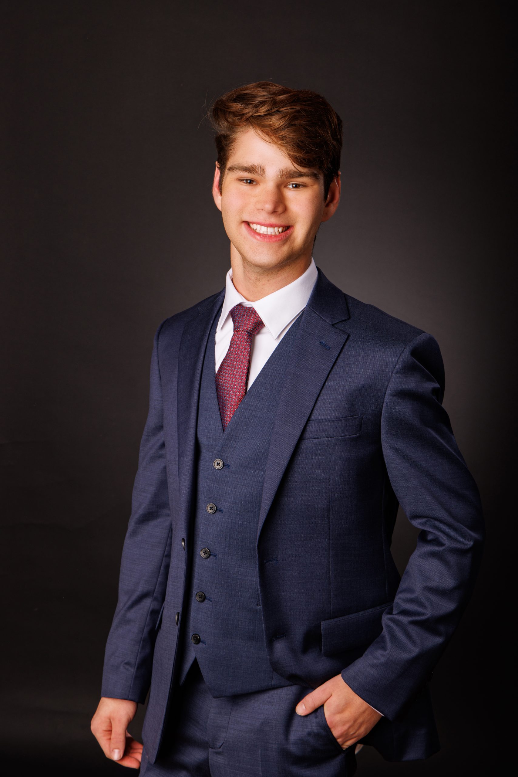 Stylishly dressed young man in a navy three-piece suit posing confidently against a dark backdrop.