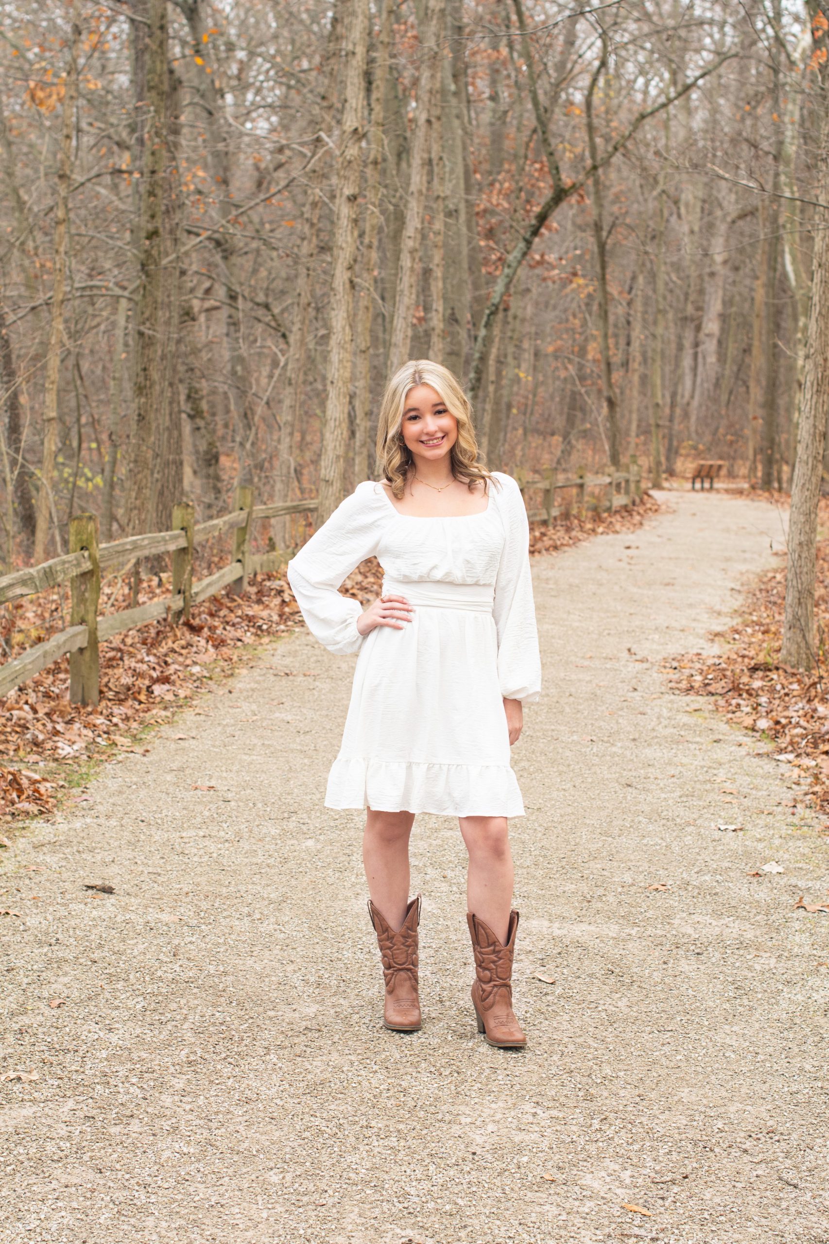 senior girl smiling on a wooded path with fallen leaves, wearing white dress and cowboy boots
